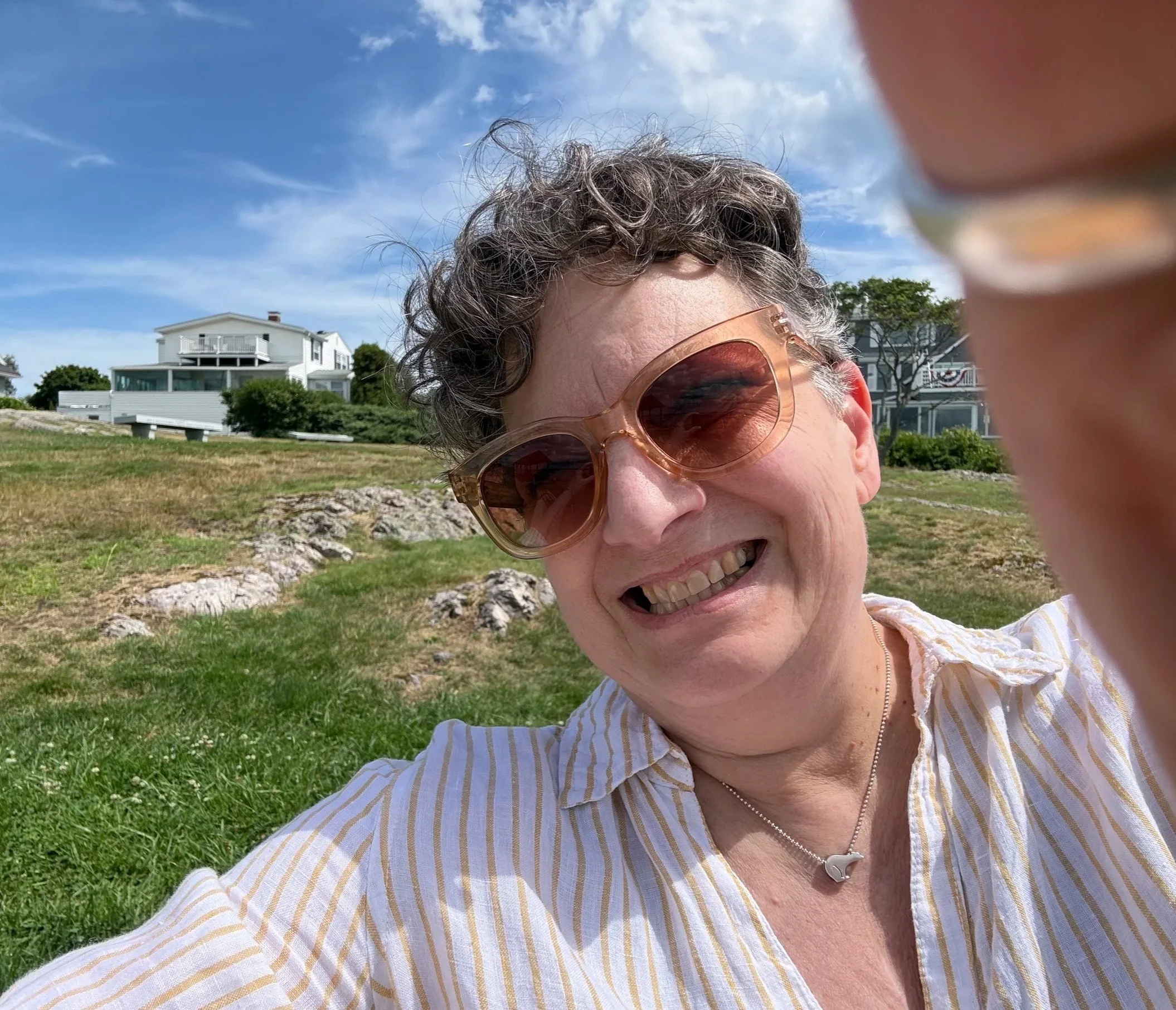 Close-up of a smiling woman with short curly hair wearing sunglasses and a striped shirt outdoors on a sunny day, with houses and trees in the background.