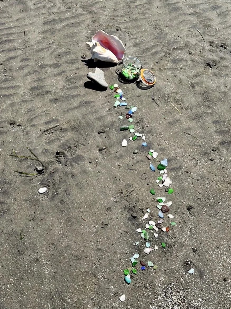 Colorful glass pieces and seashell on sandy beach, with open jar of green crystals or beads nearby.