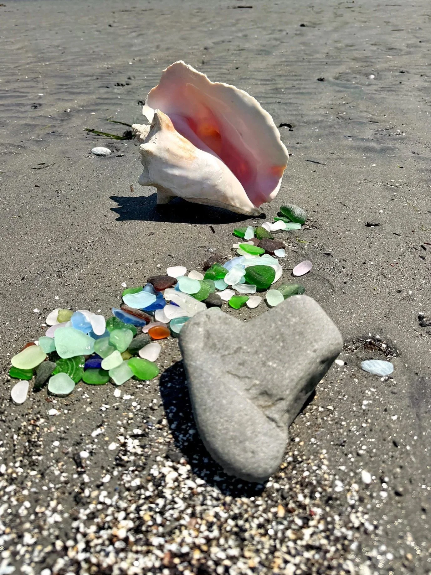 A seashell on the sandy beach surrounded by colorful sea glass and small stones, with a large heart-shaped stone in the foreground.