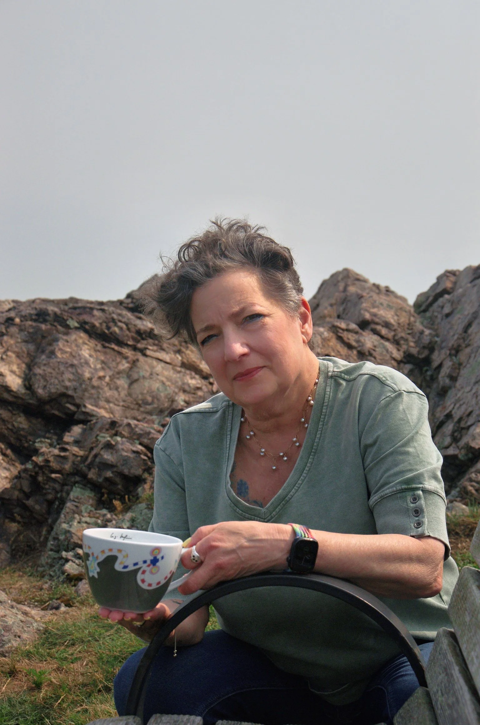 A woman with short gray hair sitting on rocks outdoors, holding a colorful mug, wearing a light green shirt, jewelry, and a smartwatch, with a landscape of rocks and sky in the background.