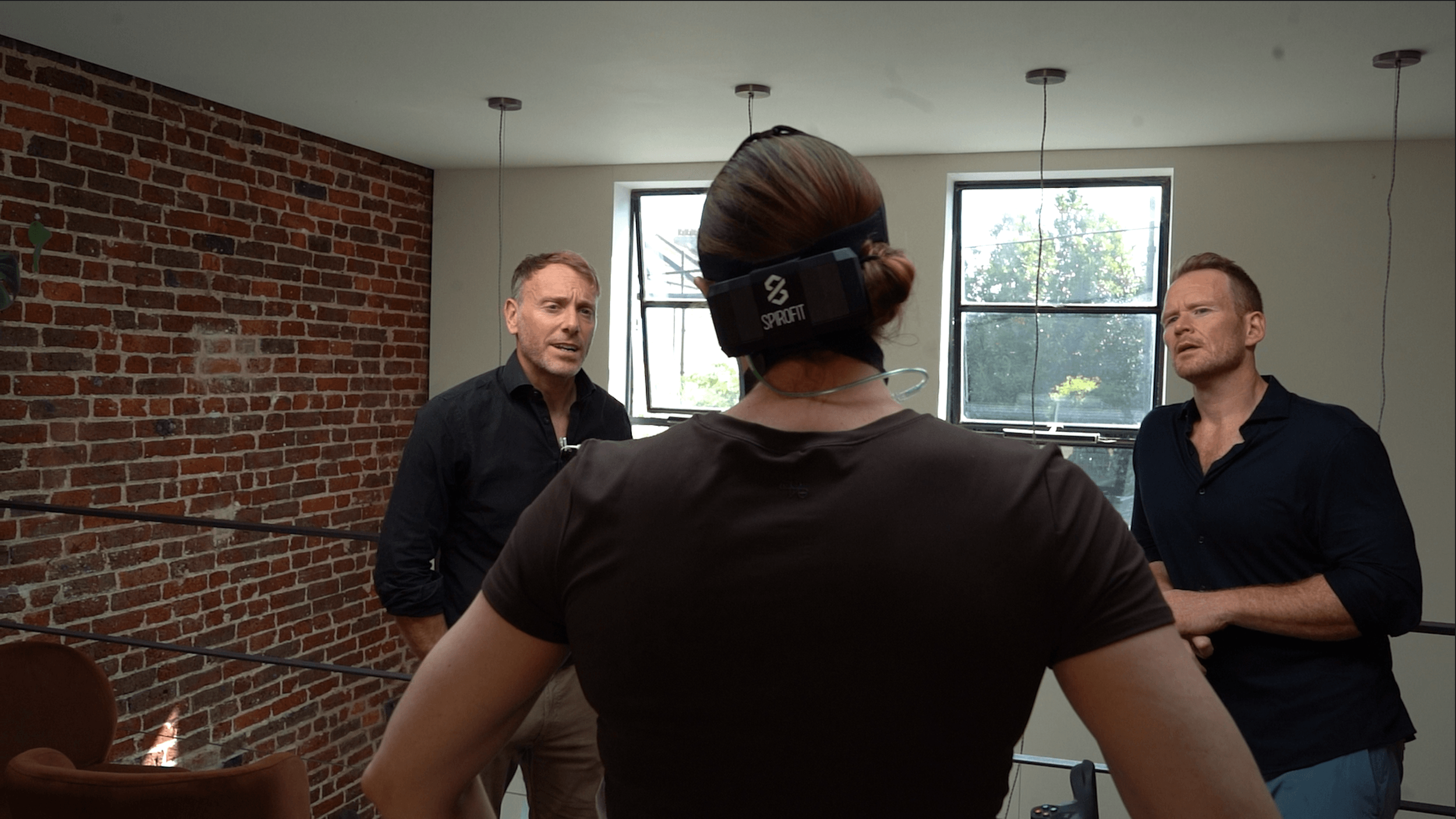 Three people engaged in conversation in a room with brick and white walls, and large windows, with a woman wearing a virtual reality headset.
