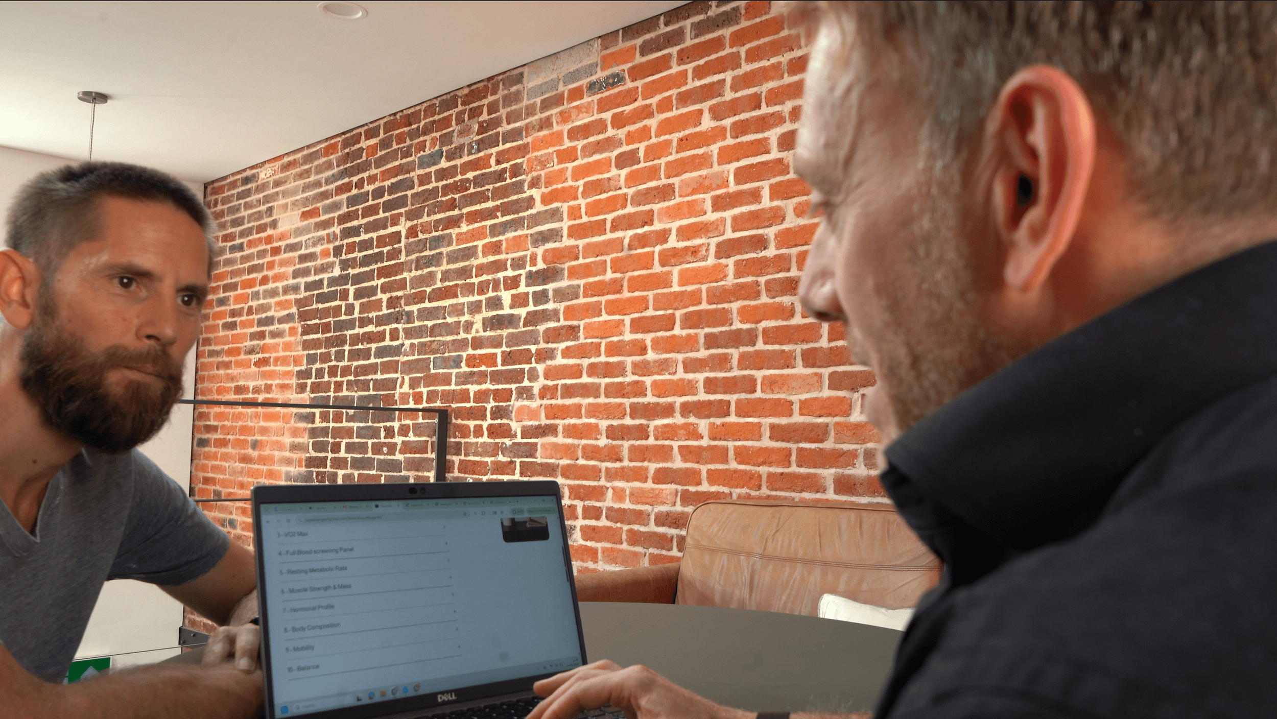 Two men sitting at a table with a laptop, in a room with a red brick wall, having a serious discussion.