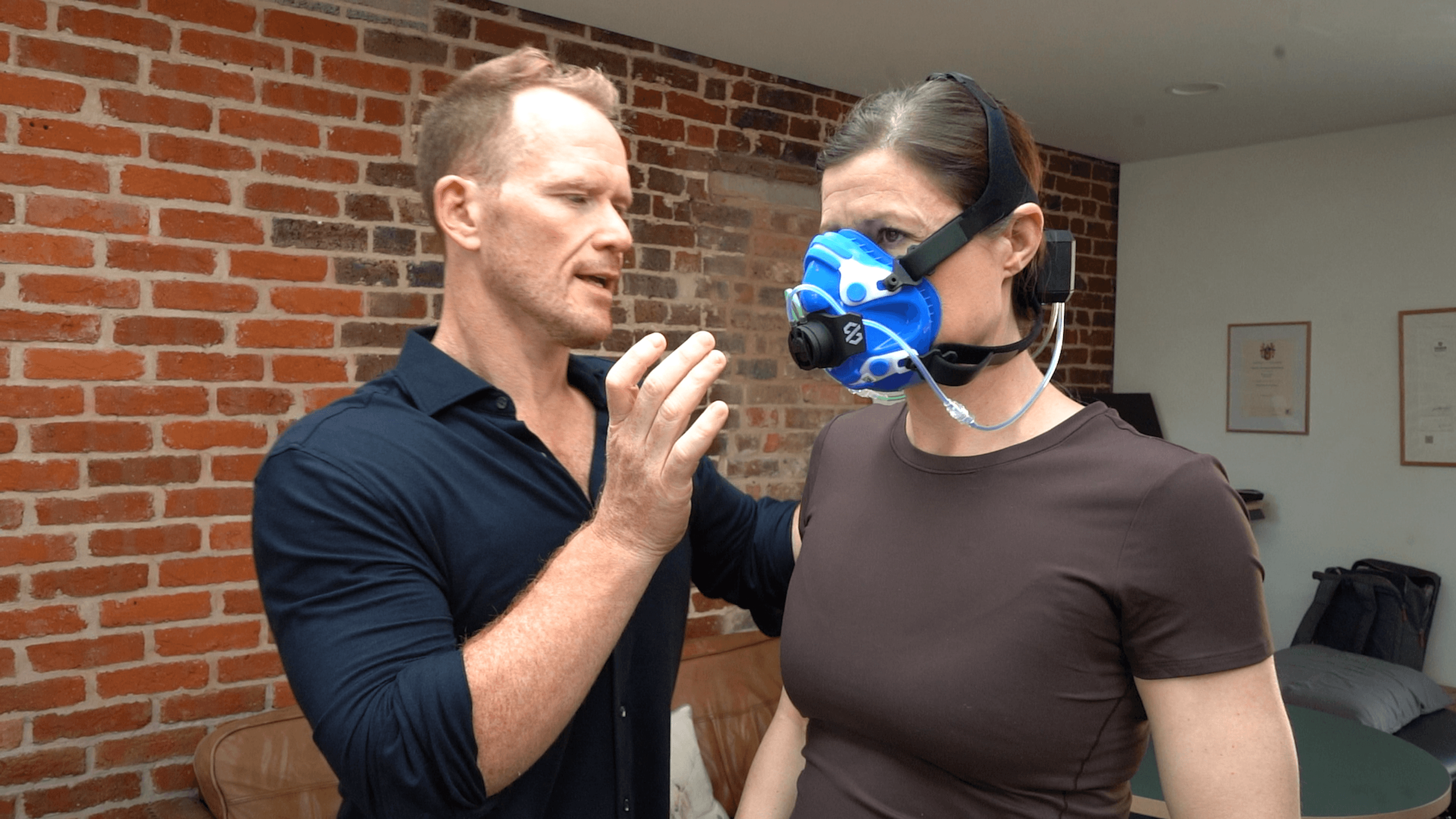 A man is adjusting a protective mask worn by a woman, who is standing with her arm at her side. They are in a room with a brick wall background, and the woman is wearing a dark shirt and headband. The man is wearing a dark shirt and has short hair.
