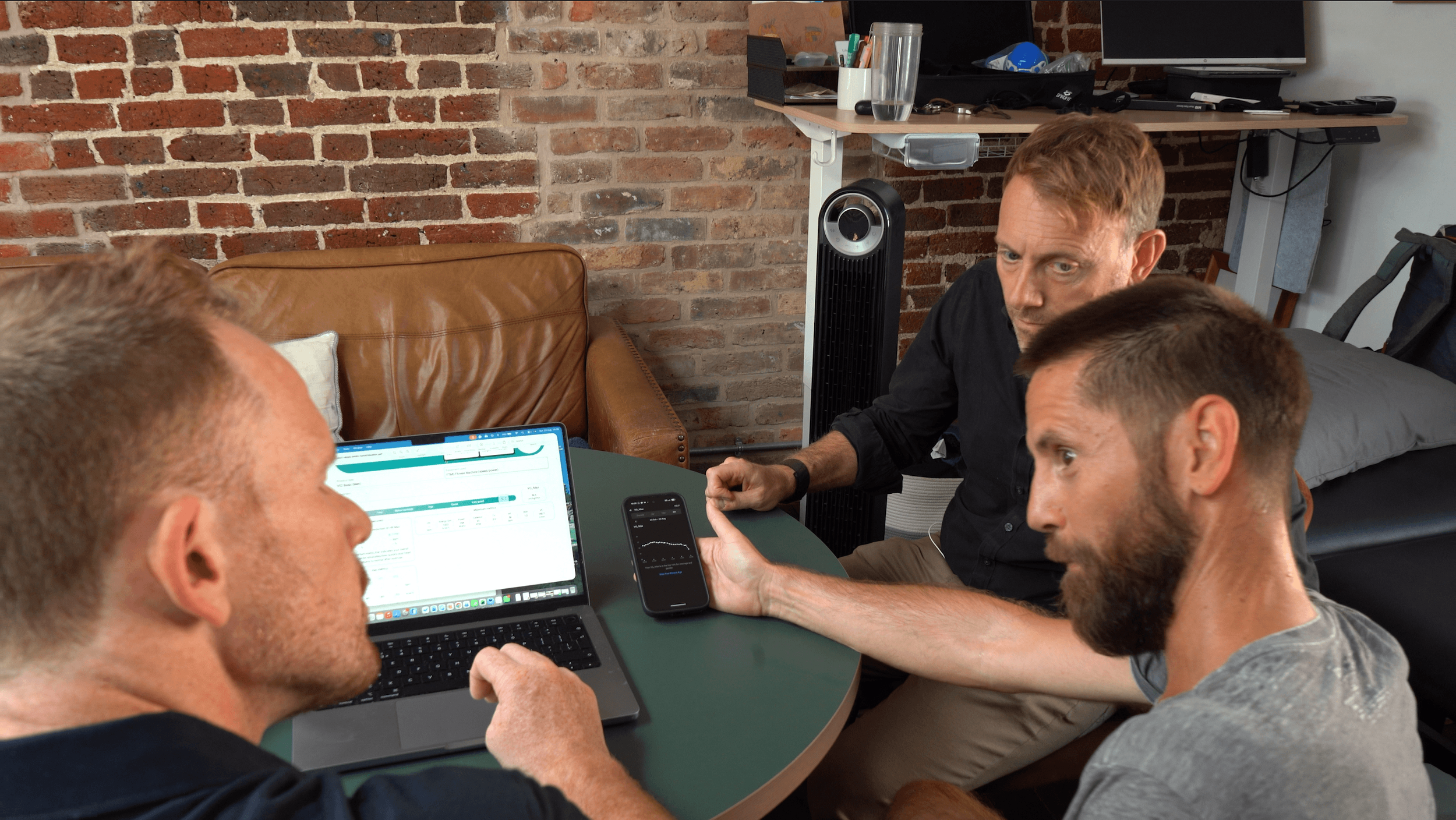 Four men having a meeting in a room with brick walls, using a laptop, smartphone, and discussing a financial chart.