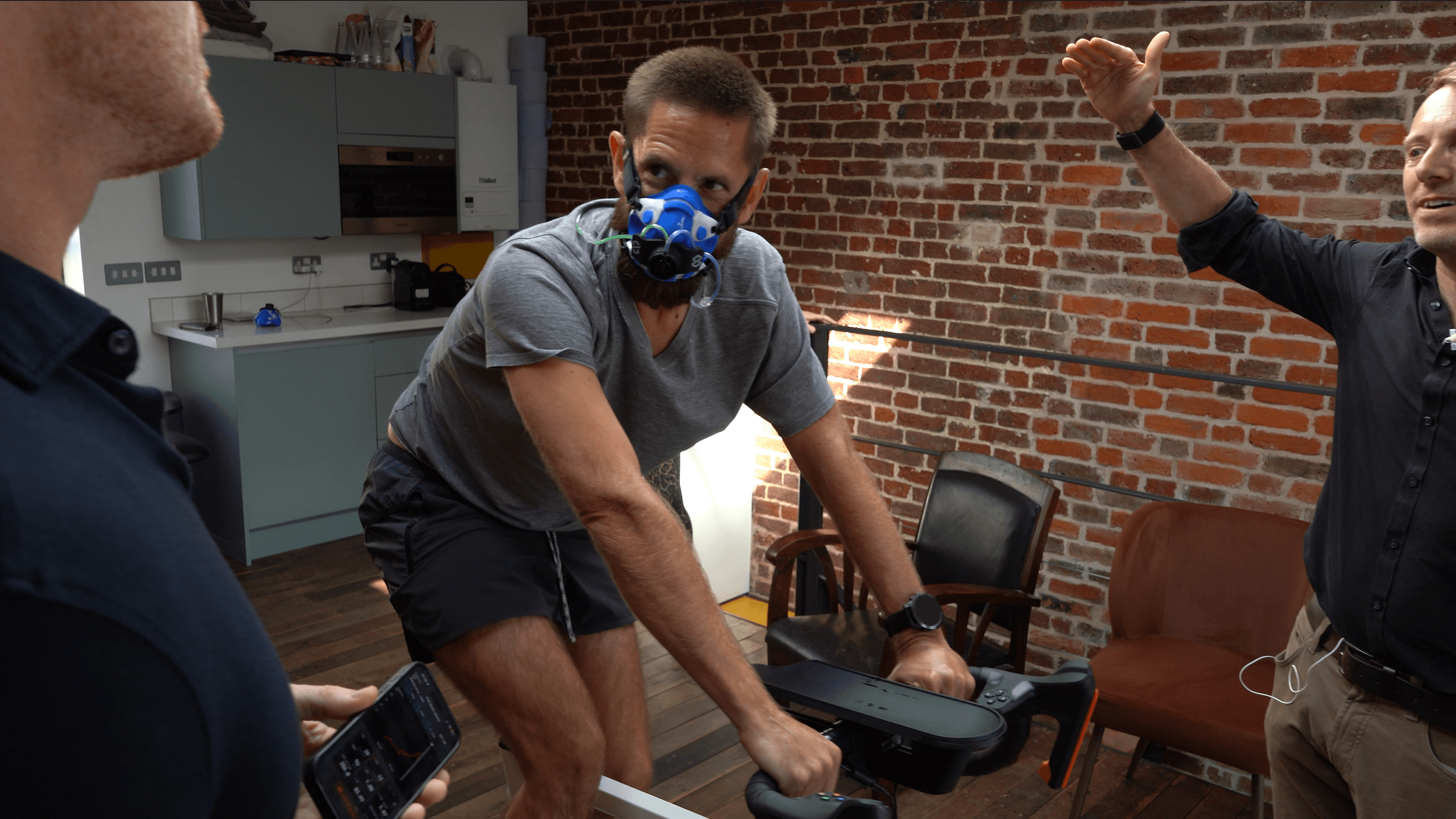 A group of men in a room with a brick wall, participating in a fitness or health-related activity, with exercise equipment, and one man wearing an air pollution mask.