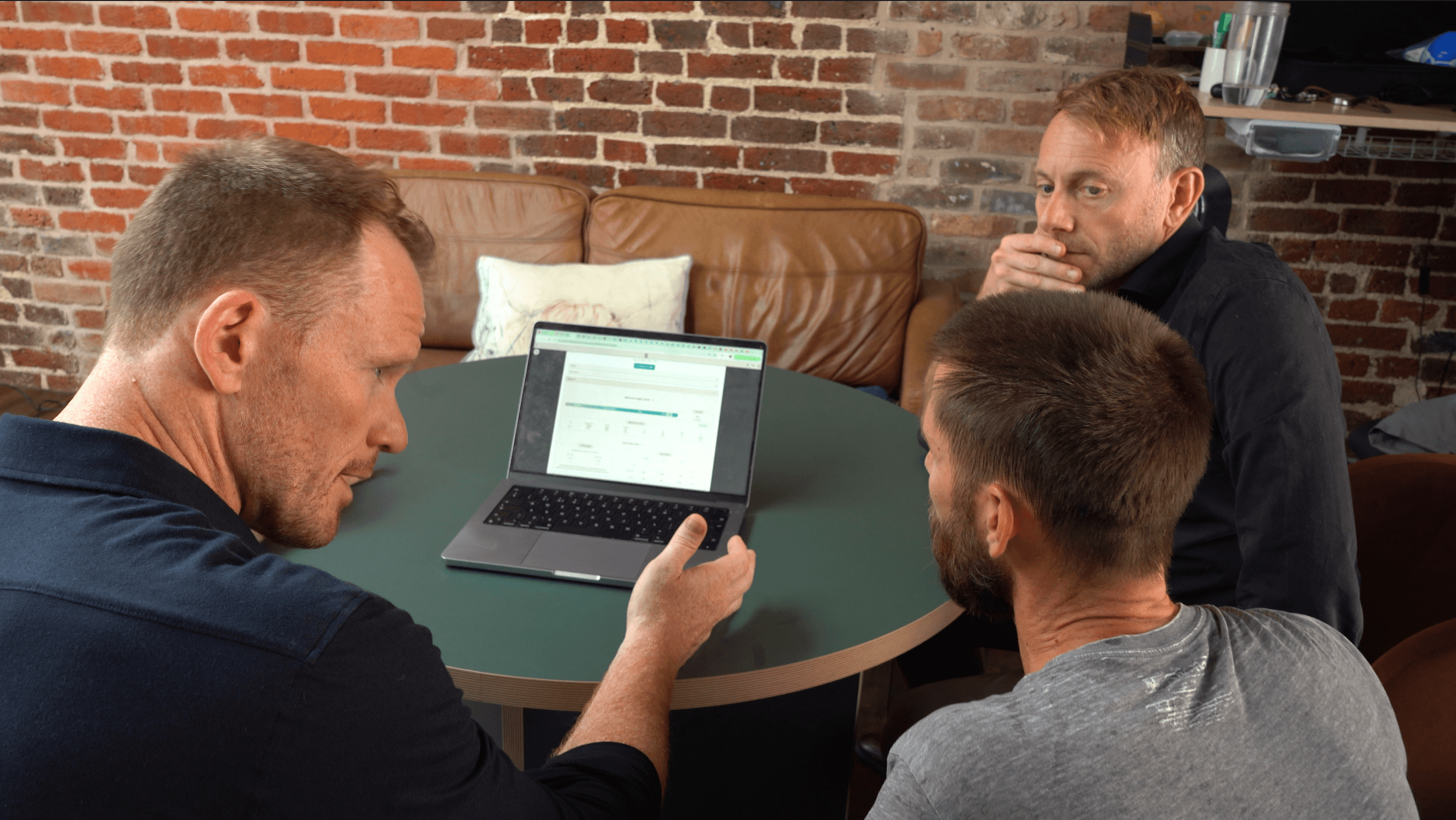 Four men sitting around a table, looking at a laptop screen. The background features a brick wall and a shelf with various items.