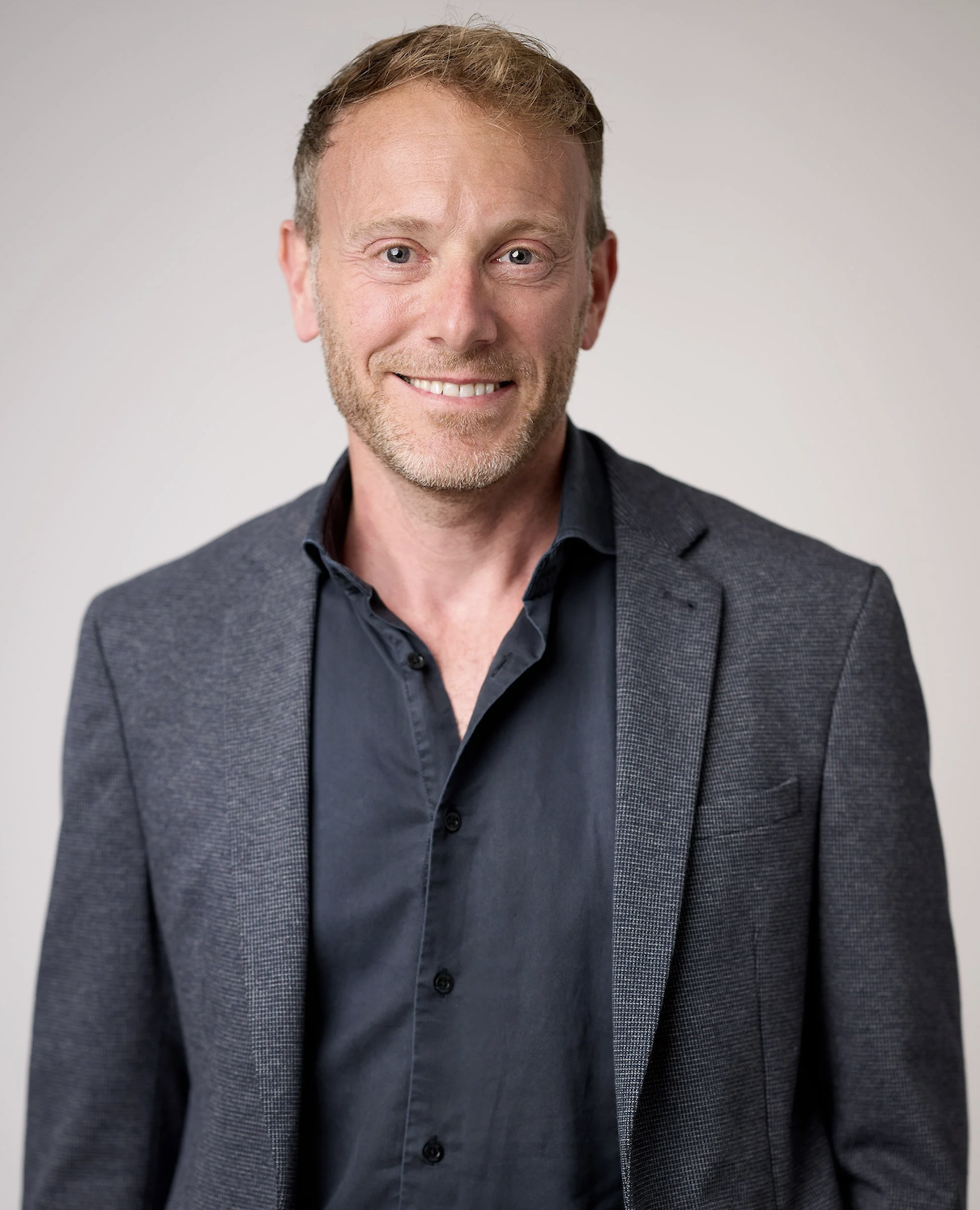 A man with light brown hair and a beard, smiling, wearing a dark shirt and a gray blazer, against a plain white background.