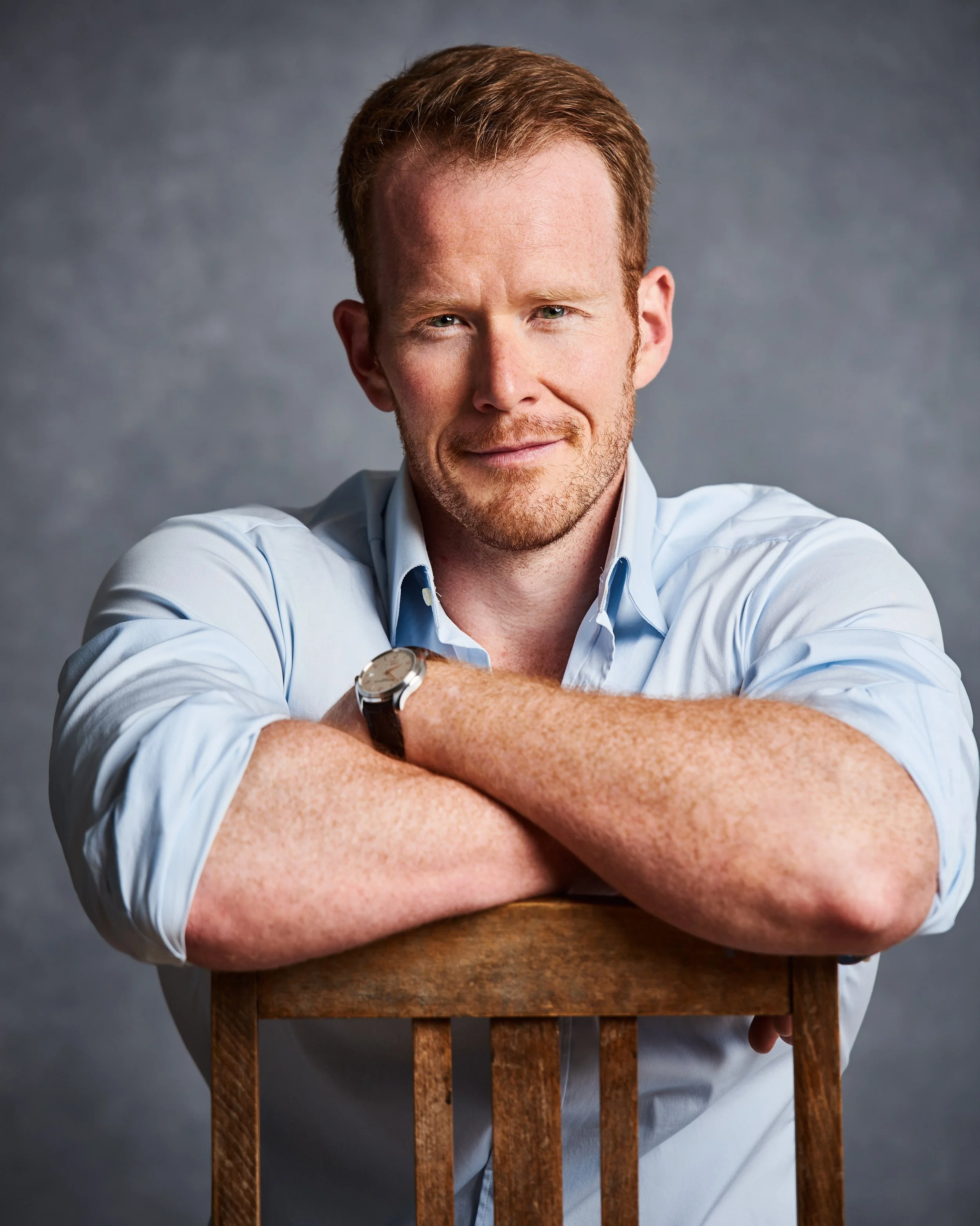 A smiling man with red hair and a beard, wearing a light blue shirt, leaning on a wooden chair with his arms crossed, against a gray background.