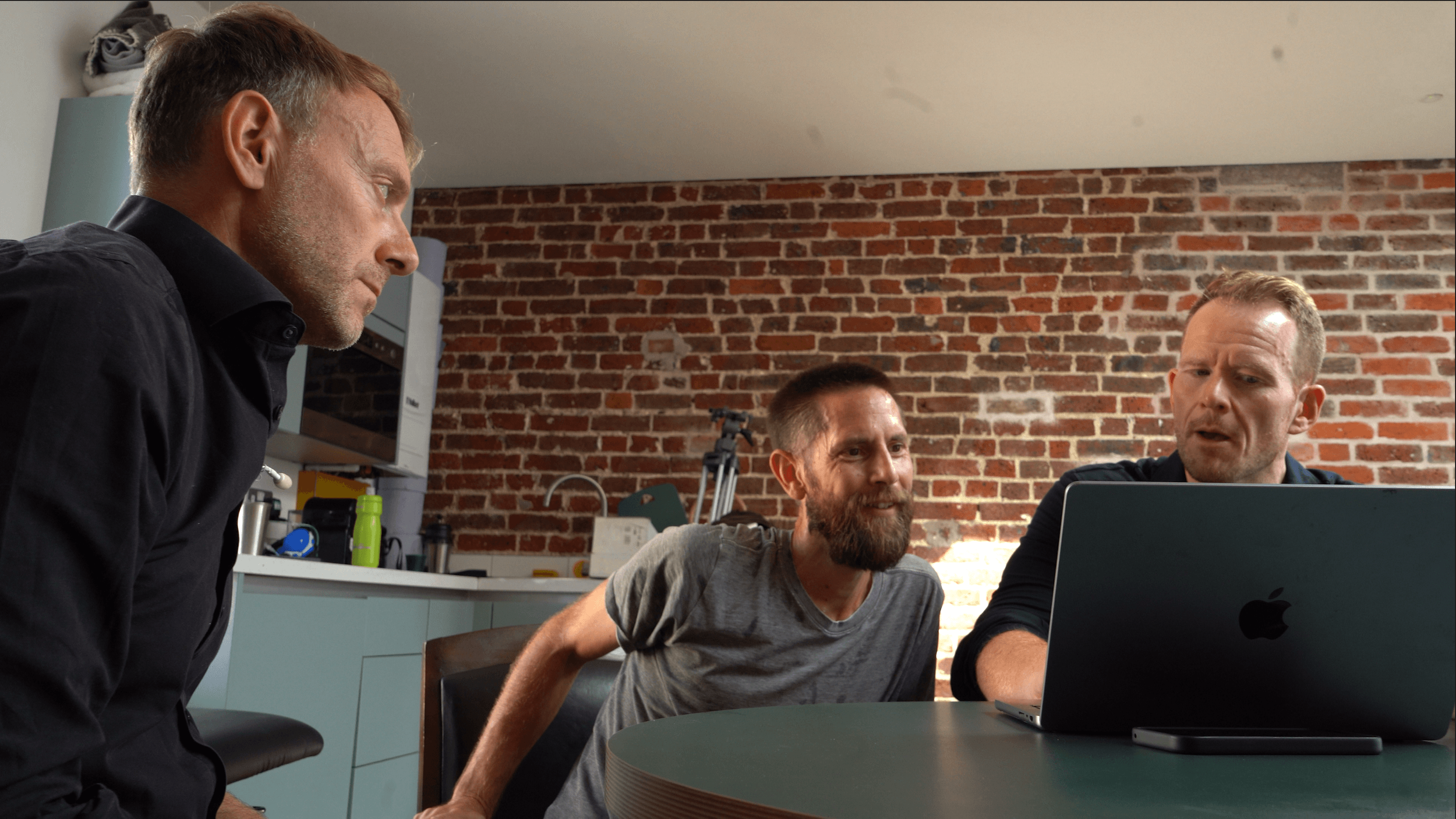 Three men gathered around a laptop in a room with a brick wall, engaged in a discussion or collaboration.