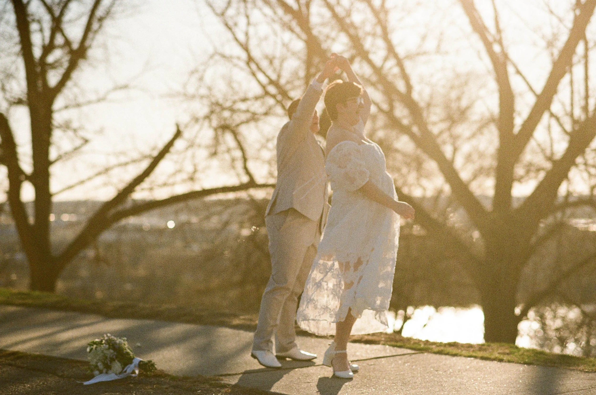 Two women dressed in wedding attire dance outdoors during golden hour, while holding hands, with bare trees and warm sunlight in the background.