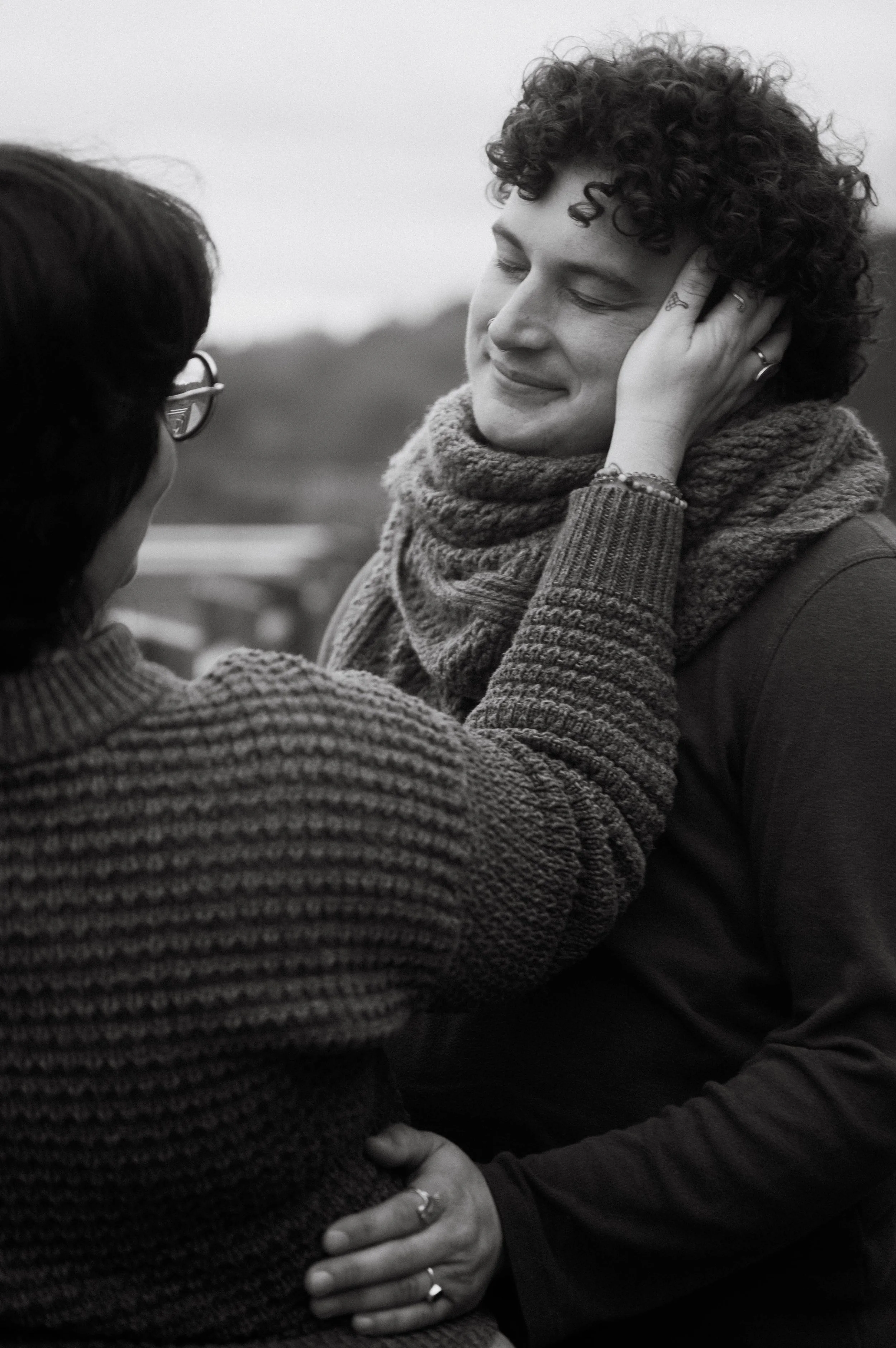 A black and white photo of two people hugging. One person, with curly hair and a knitted sweater, is smiling with their eyes closed, holding the other person gently. The other person is wearing glasses and a textured sweater, with their face partially visible.