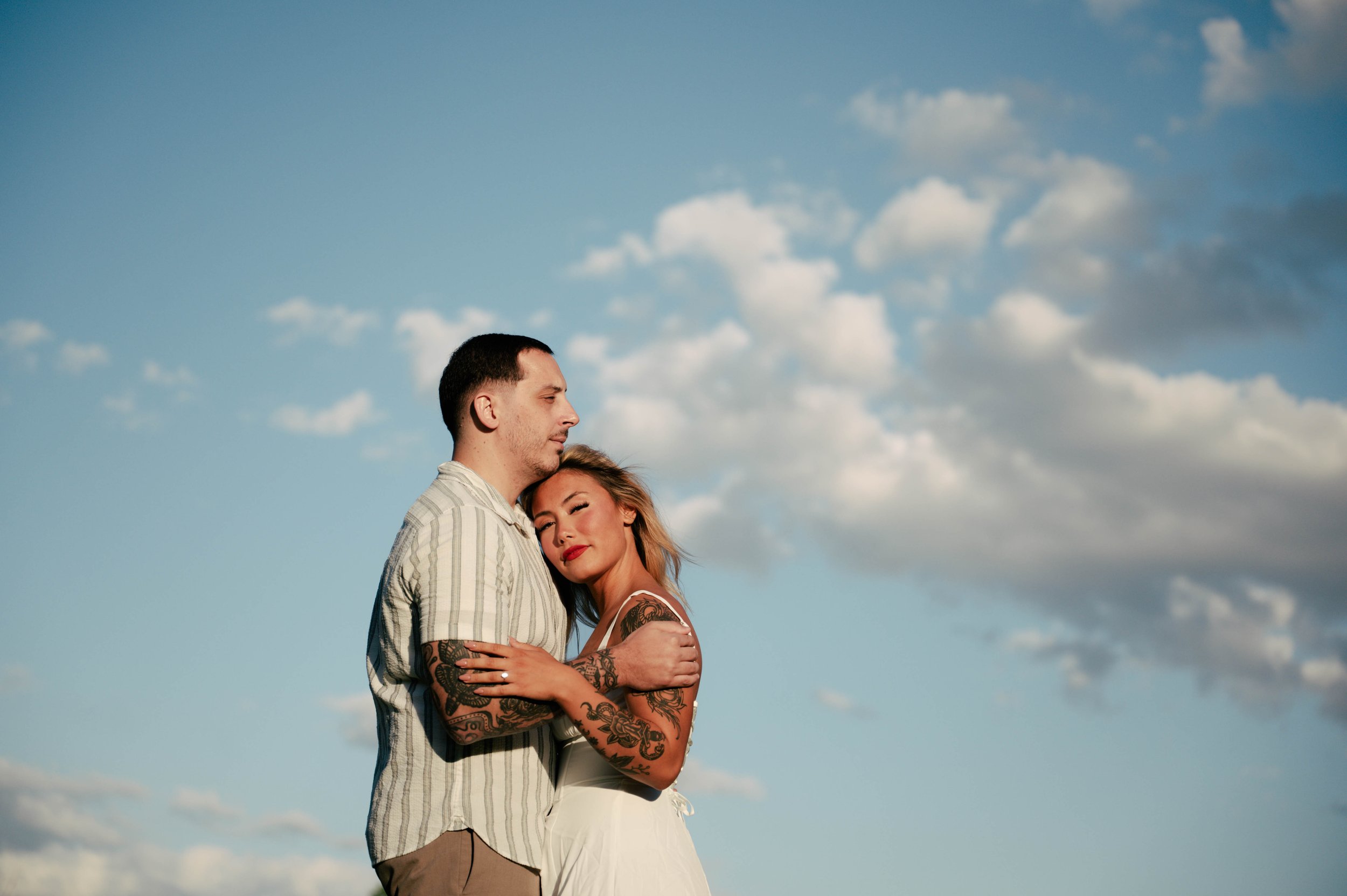 A man and a woman embrace outdoors against a bright blue sky with scattered clouds. The man has short dark hair, a light beard, and is wearing a striped shirt. The woman has blonde hair, tattoos on her arms, and is wearing a white dress with spaghetti straps. Both have their eyes closed and appear content.
