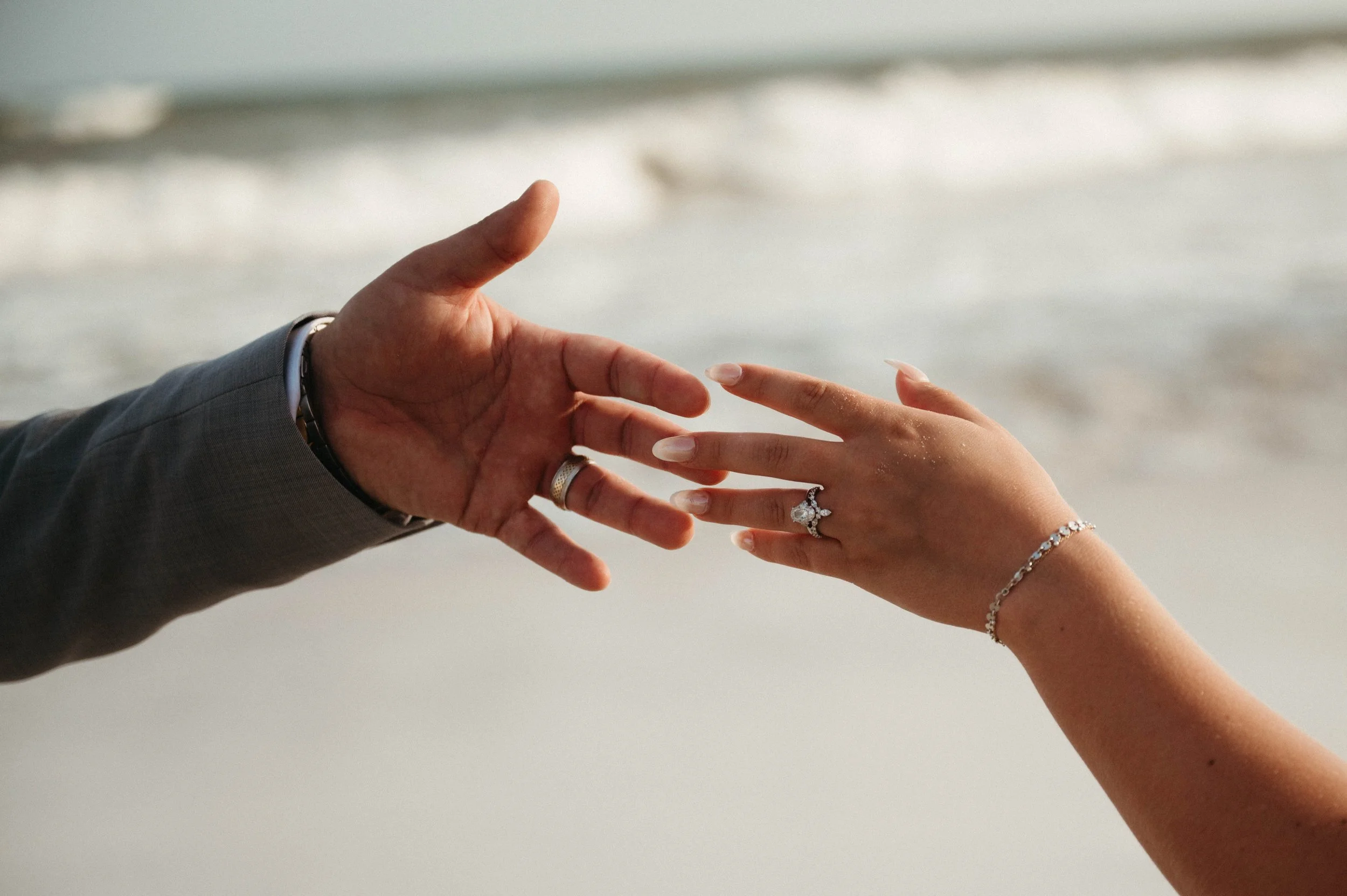 Close-up of a man and woman holding hands, the woman wearing an engagement ring and a bracelet, the man wearing a gray suit and a watch, with a blurred outdoor background.
