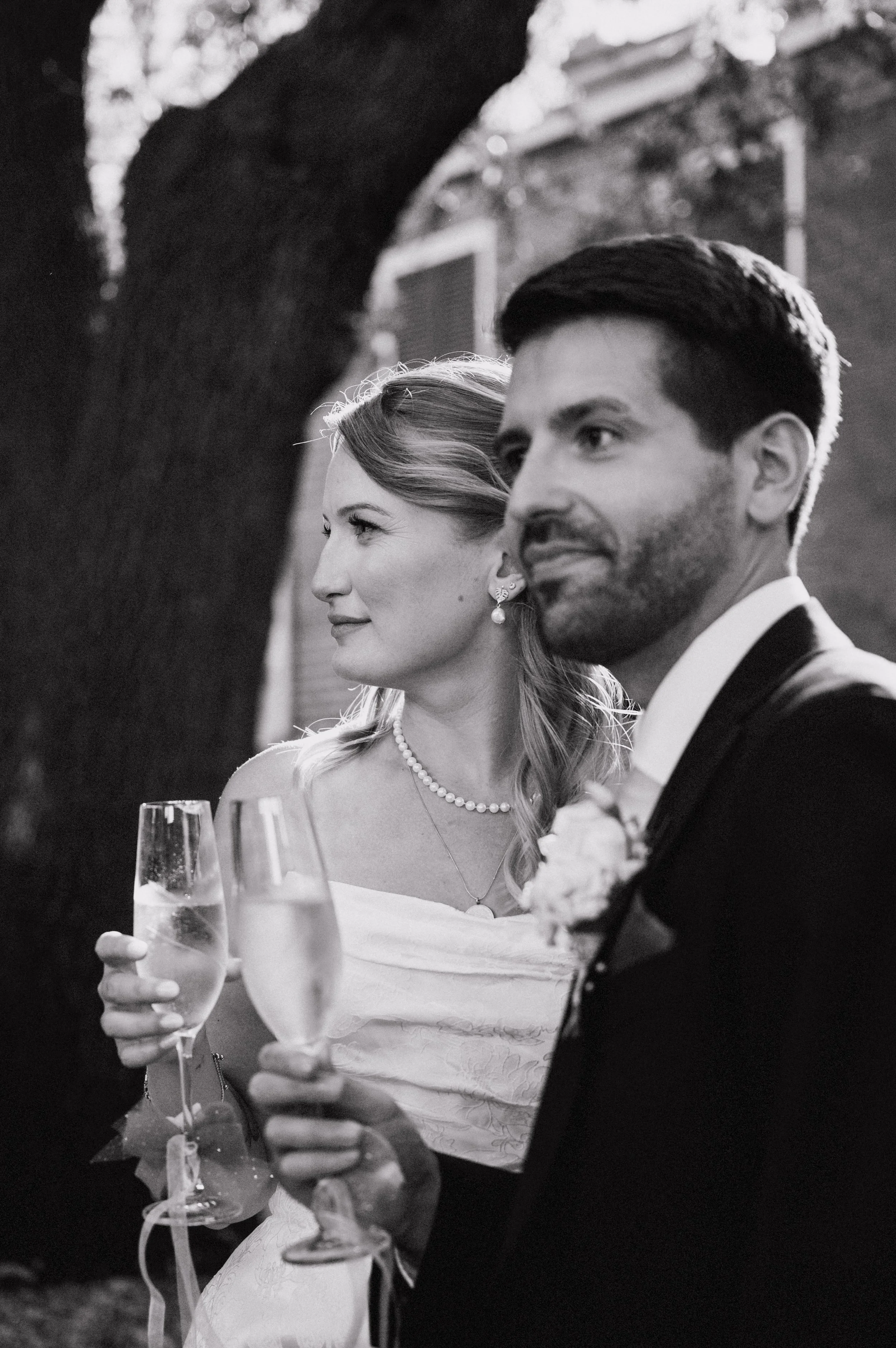 A black-and-white photo of a bride and groom holding champagne glasses during their wedding celebration. The bride is smiling and wearing a pearl necklace and earrings, while the groom is looking to the side, dressed in a tuxedo. They are standing ou
