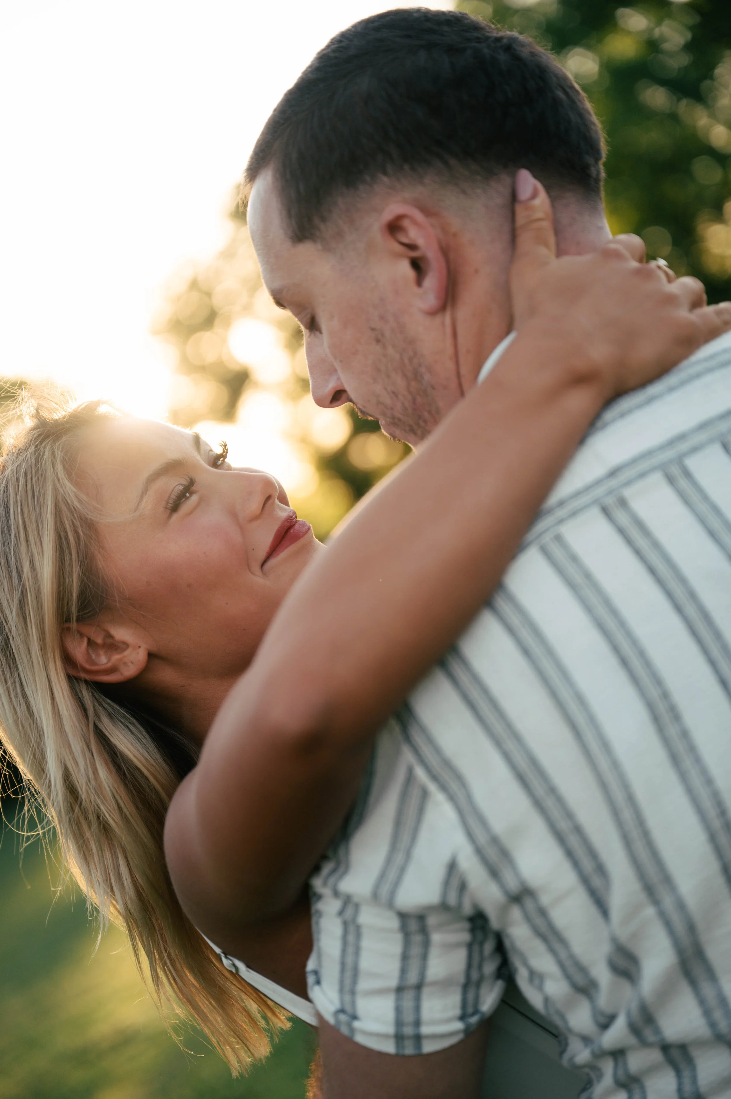 A couple sharing a close, intimate moment outdoors during sunset, with the man holding the woman around her shoulders and the woman looking up at him, both smiling.