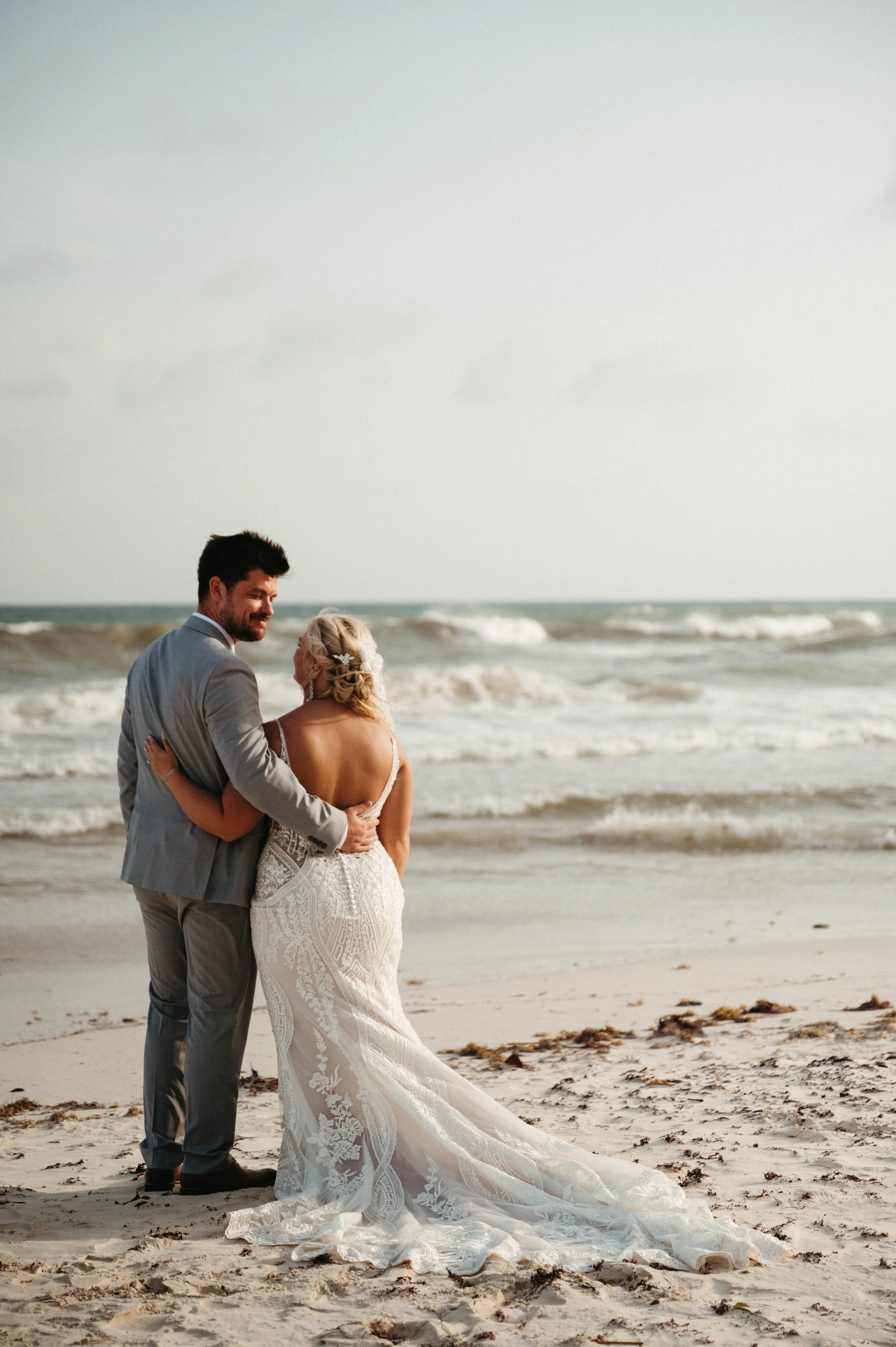 A couple in wedding attire standing on the beach, the groom in a gray suit and the bride in a white lace wedding dress, embracing each other with ocean waves in the background during daytime.