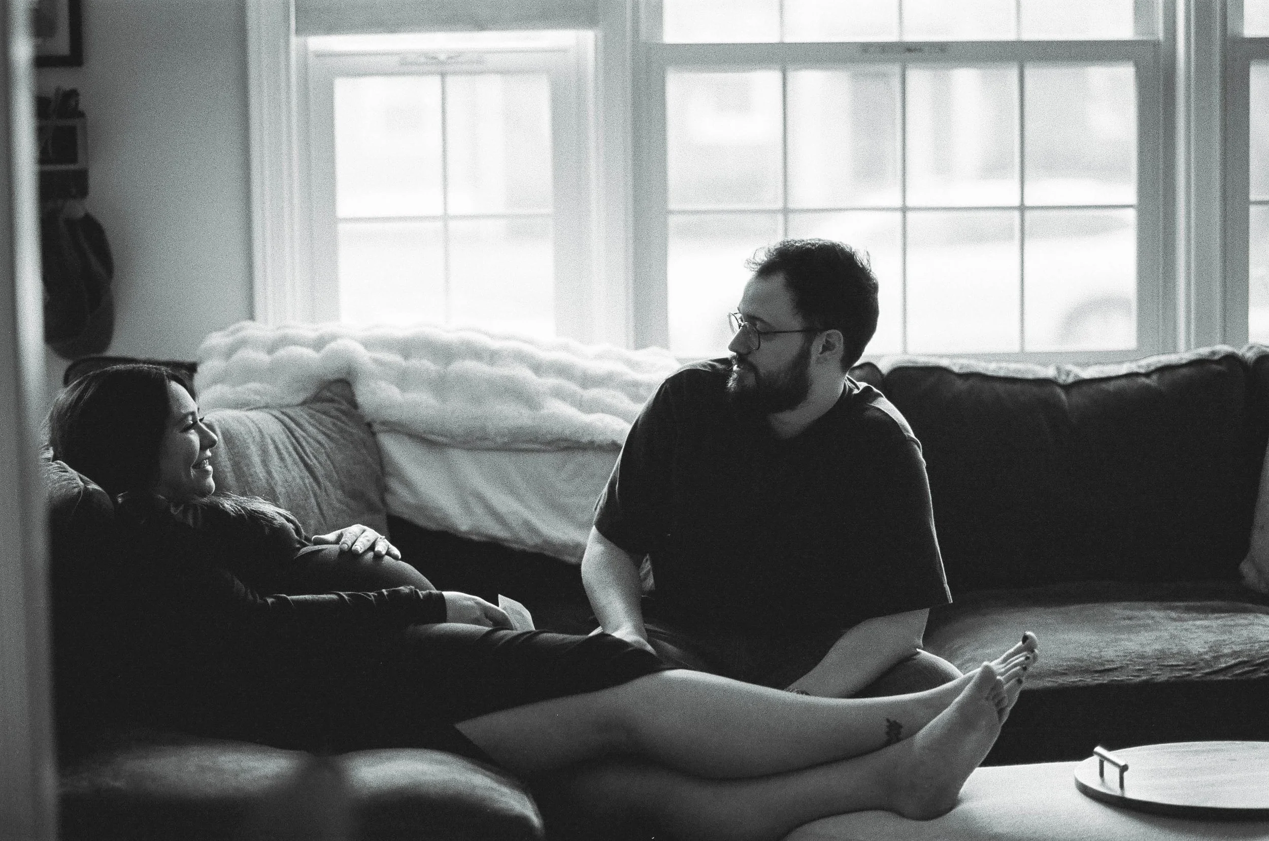 A woman lying on a couch smiling and holding her pregnant belly and looking at a man sitting beside her in a living room with large windows.