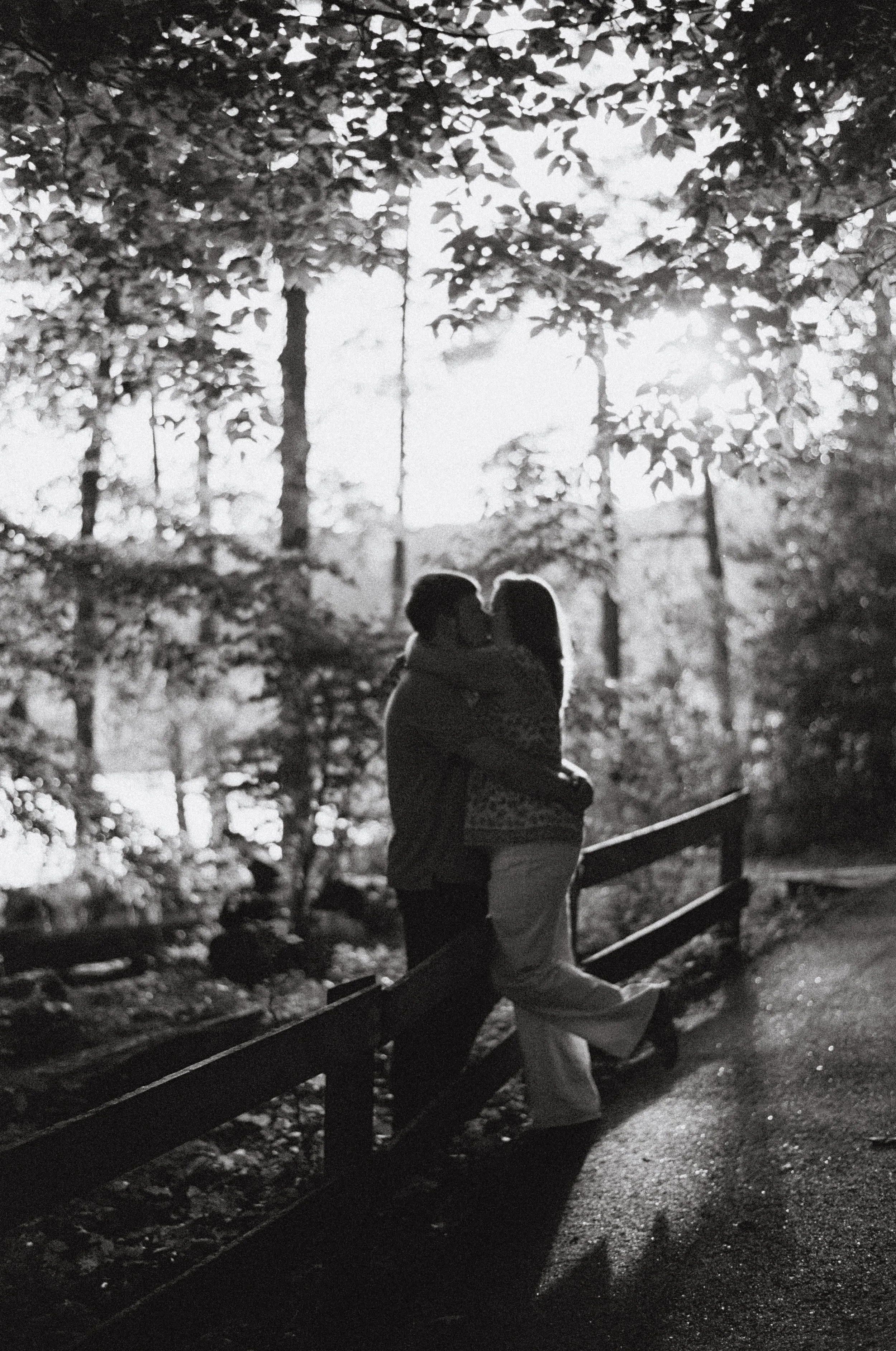 A couple kissing at a park during sunset, surrounded by trees and foliage, in black and white.