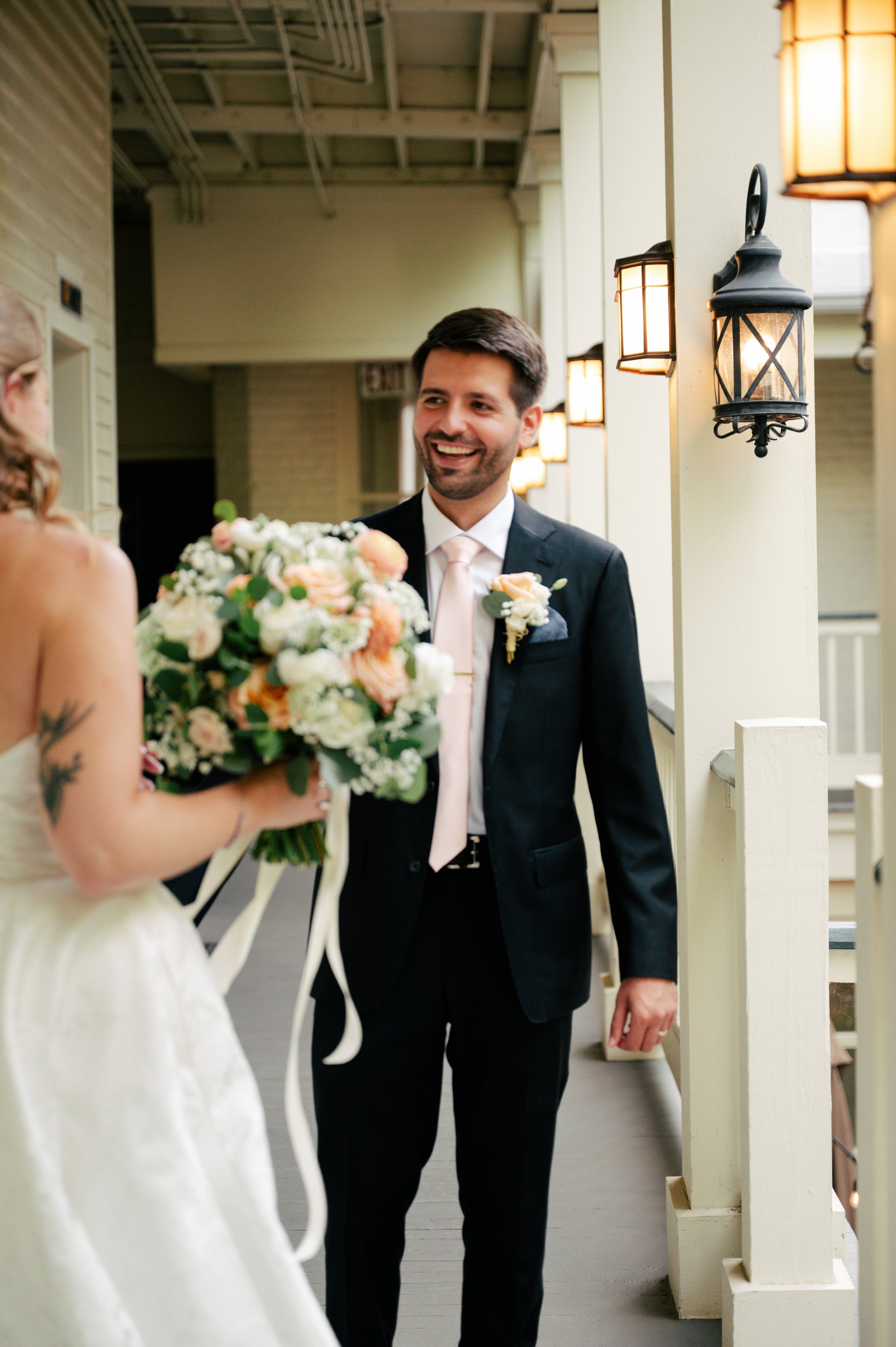 A groom in a black suit and pink tie smiling and looking at a bride holding a bouquet of white and pink flowers during a wedding ceremony.