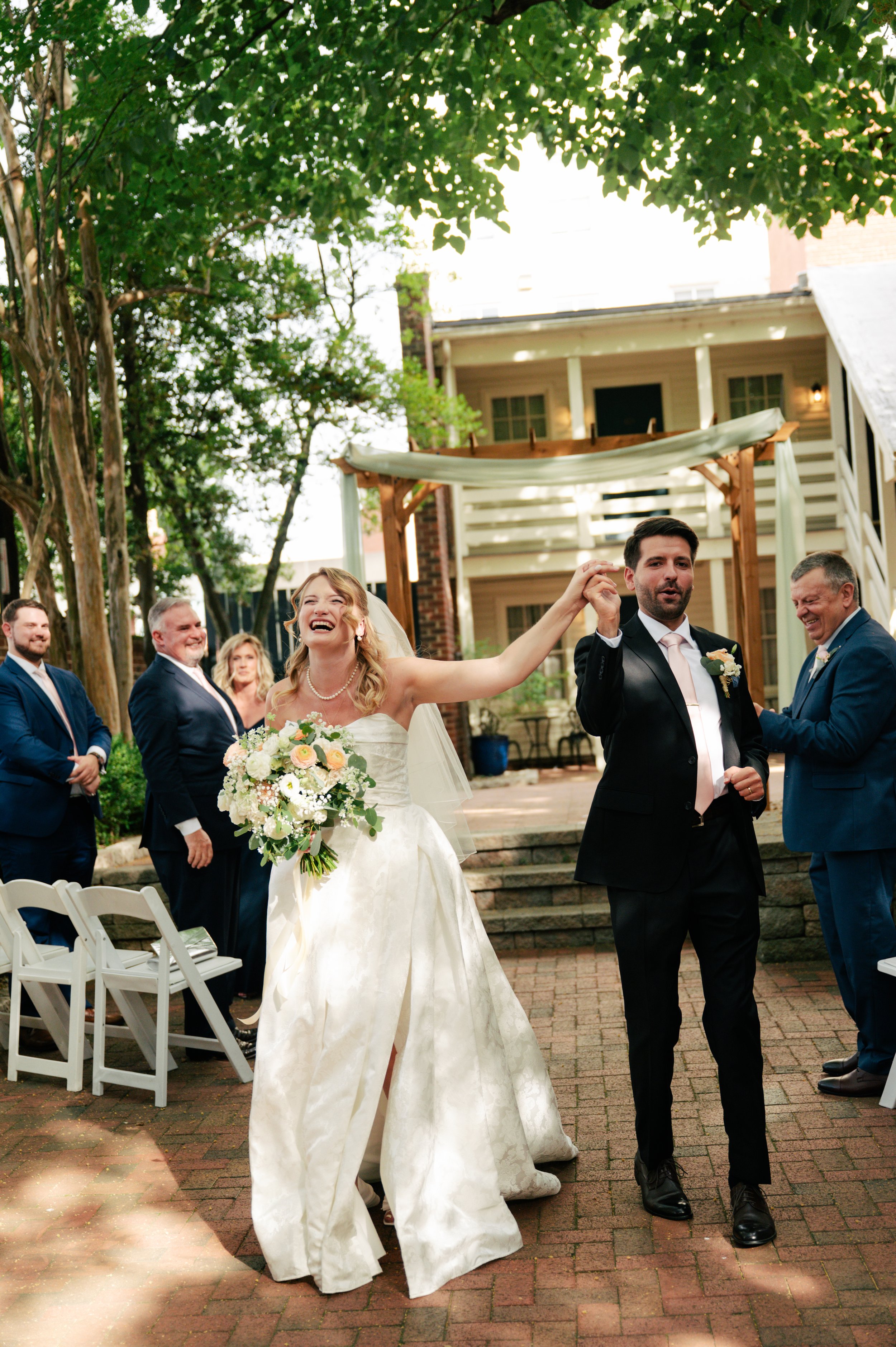 A bride with curly blonde hair in a strapless wedding gown holding a bouquet of flowers, smiling as she dances with a groom who has dark hair and a beard, dressed in a black tuxedo, at an outdoor wedding reception with friends and family in the backg