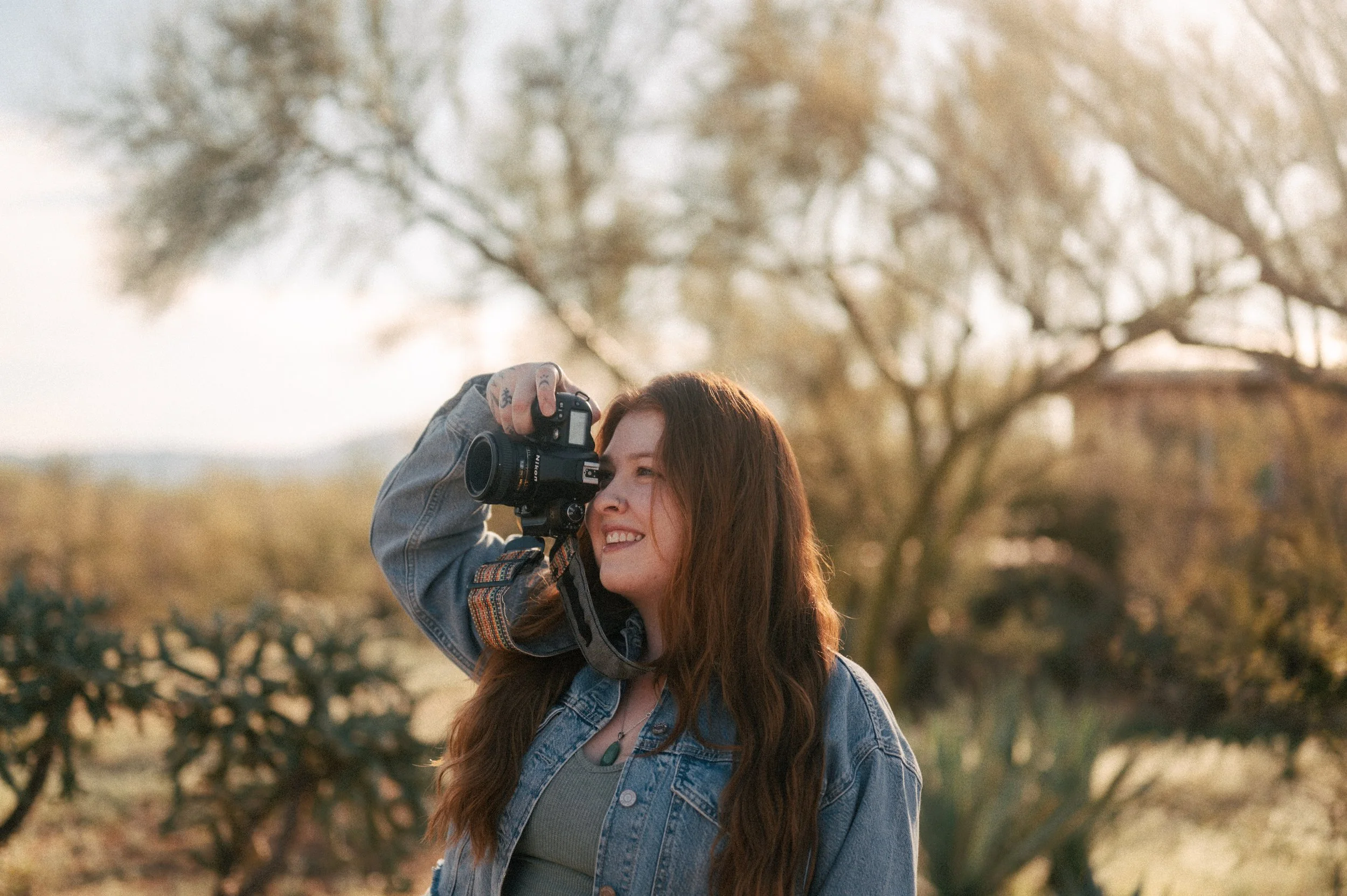 A woman with long red hair holding a camera up to her eye, smiling outdoors in a park-like setting with trees in the background during golden hour.