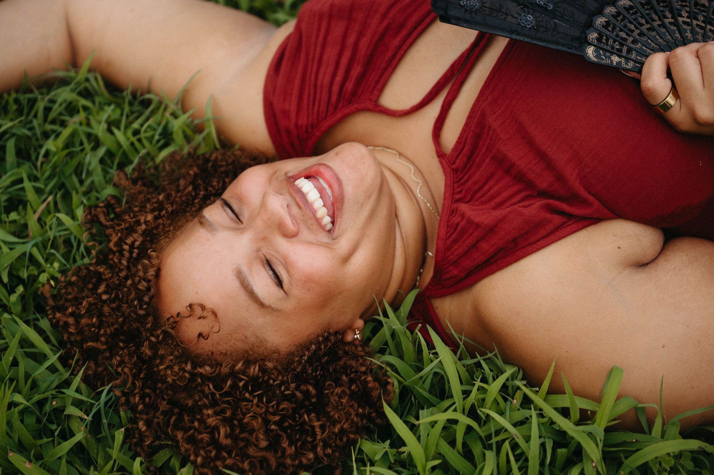 A woman with curly hair lying on grass, smiling, wearing a red top and holding a fan.