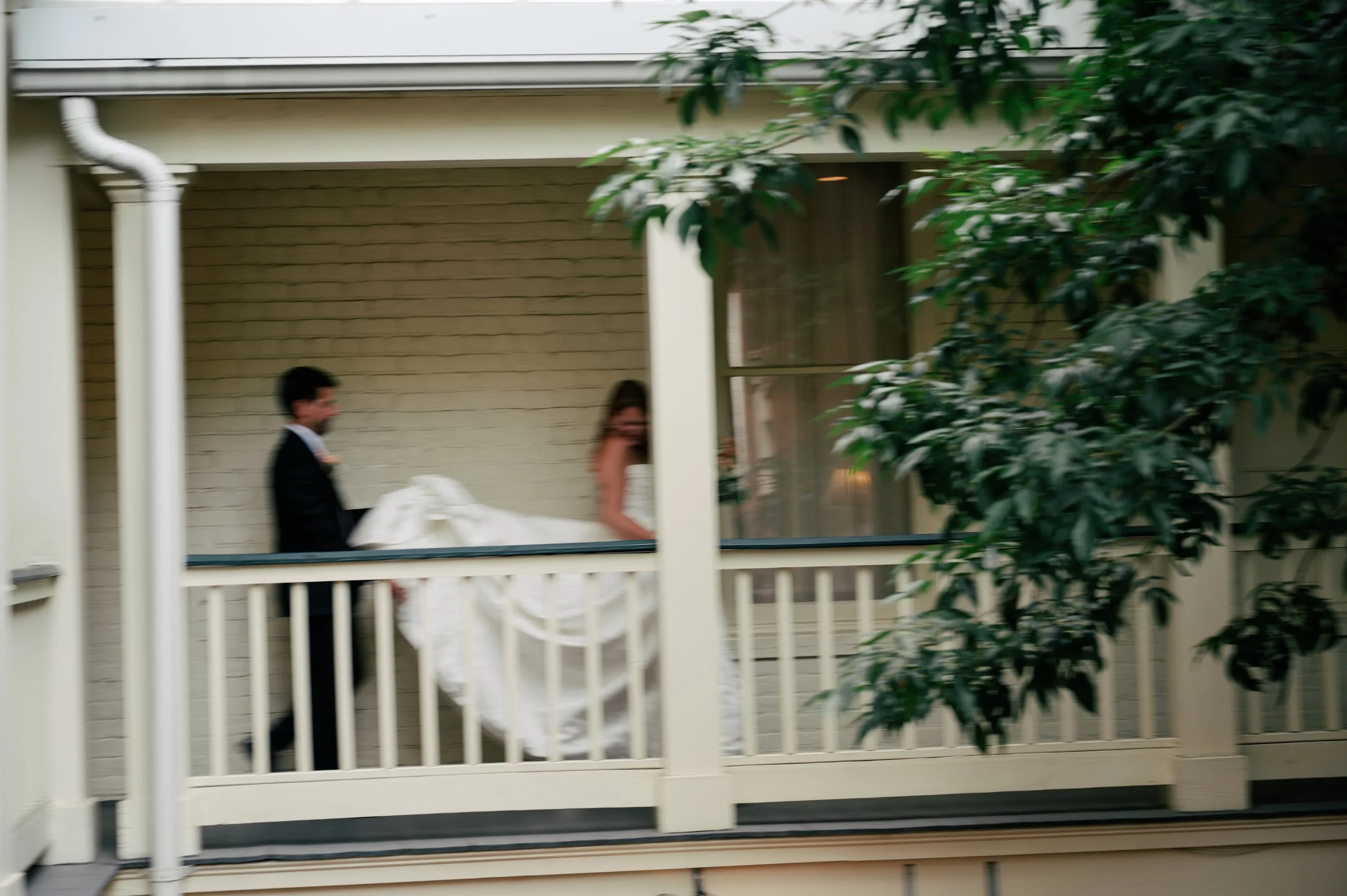 A man and woman dressed in formal attire, possibly wedding clothes, walking on a porch with white railings, with a house and trees in the background.