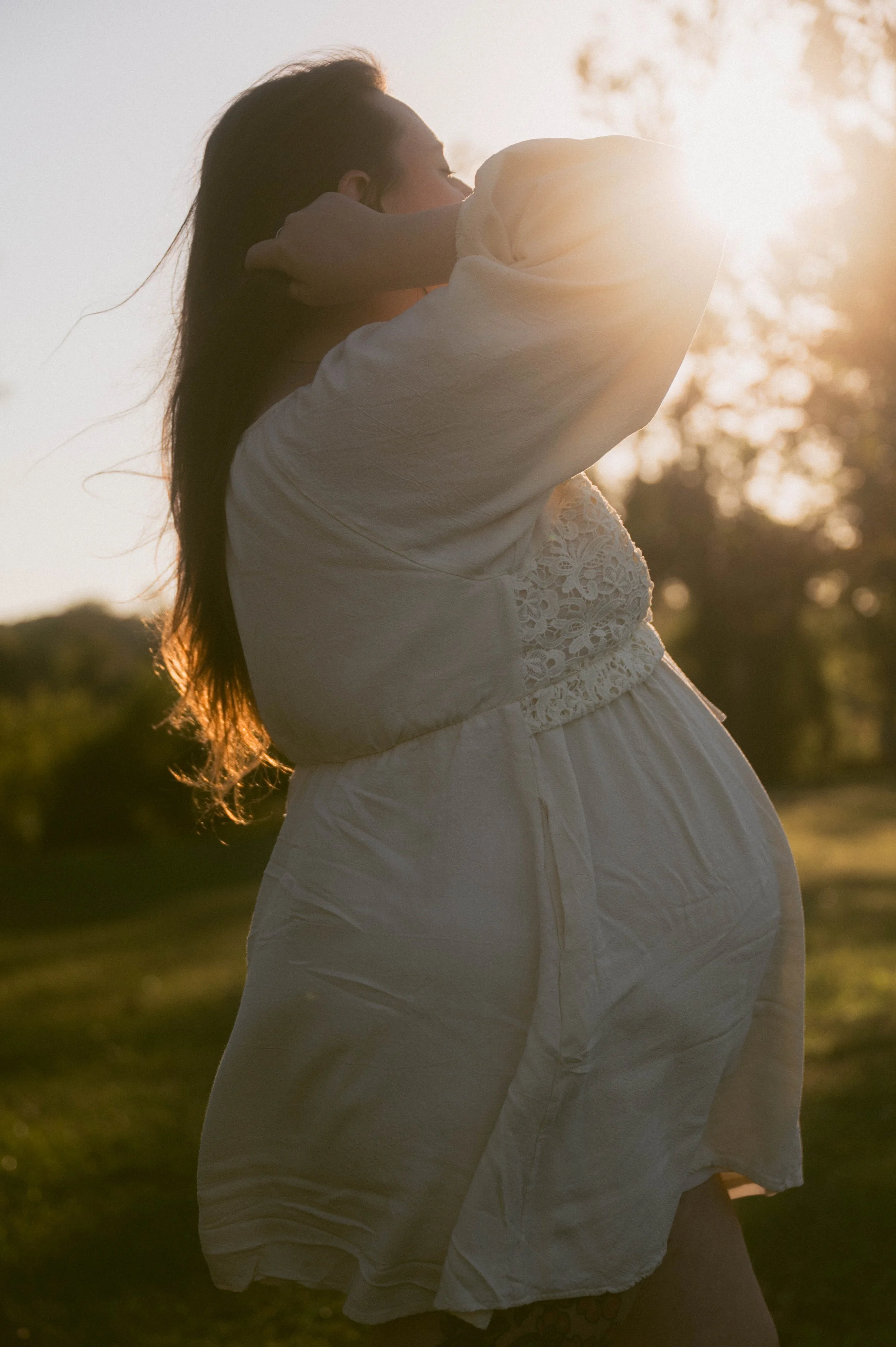 A woman in white dress embracing a pregnant stomach outdoors during sunset.