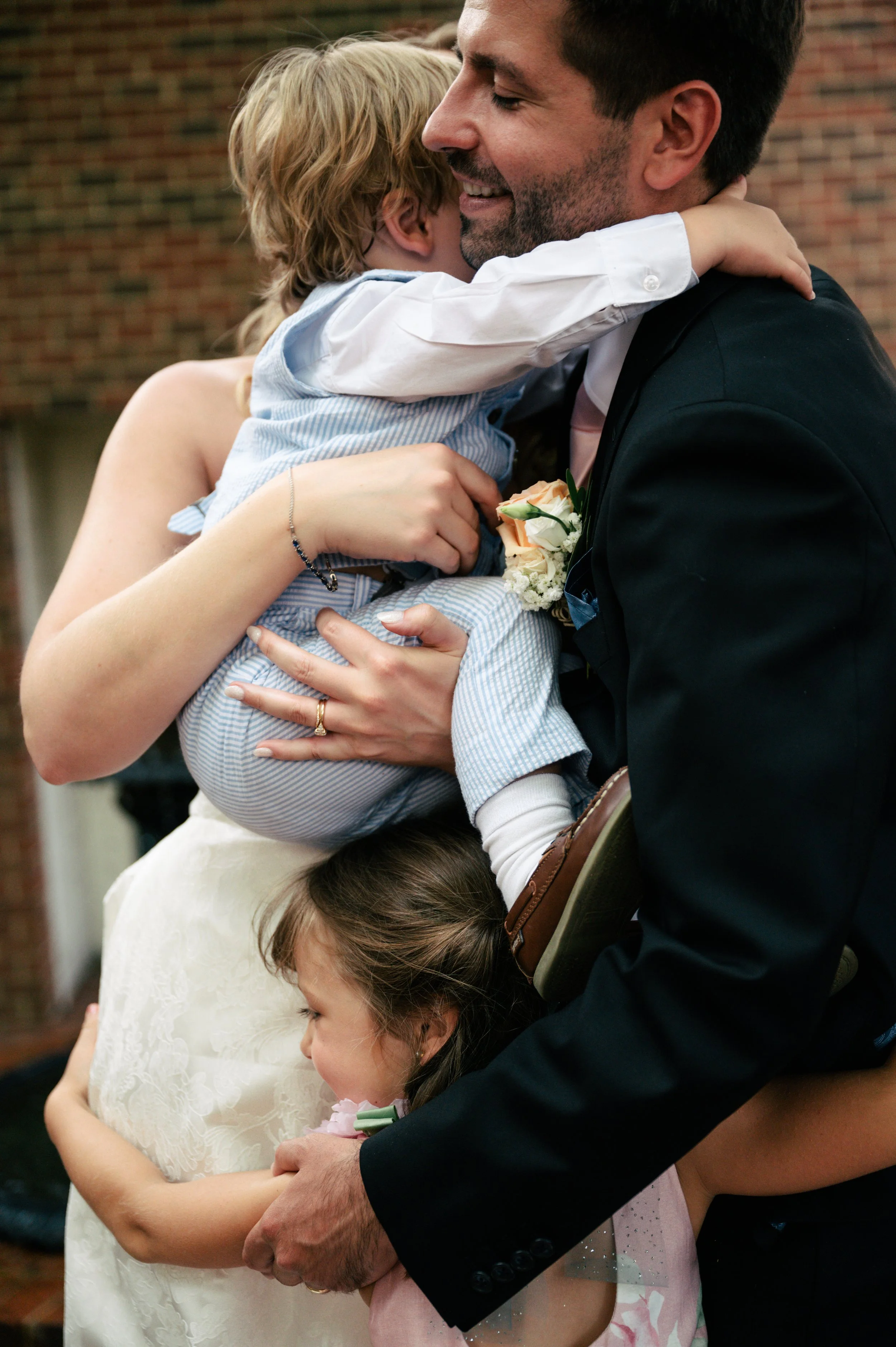 A man, woman, and two children hugging at a celebration, with the man dressed in a suit and the woman in a lace dress.