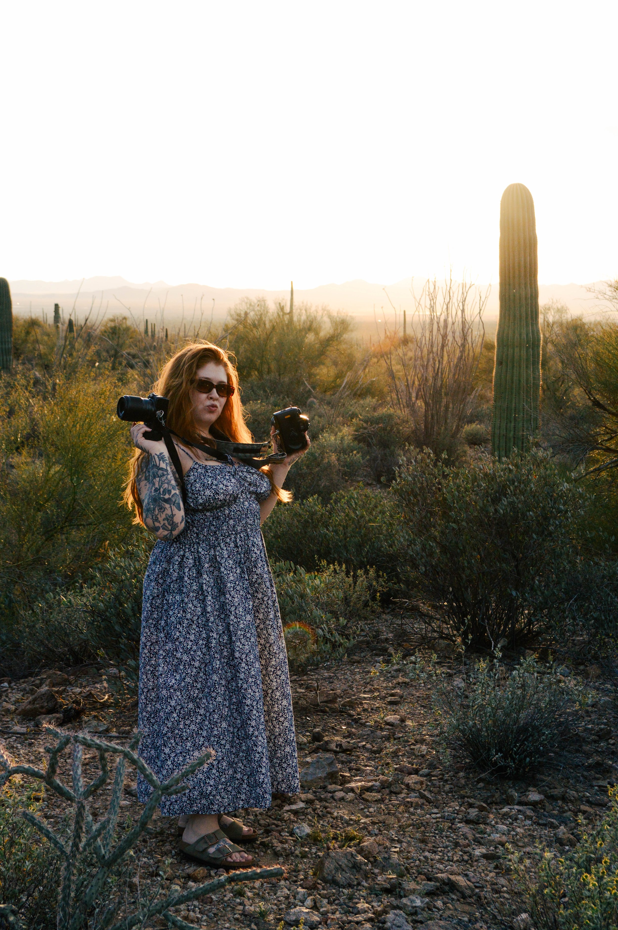 A woman with long red hair, wearing sunglasses, a floral dress, and sandals, stands in a desert landscape at sunset holding a camera in her right hand.