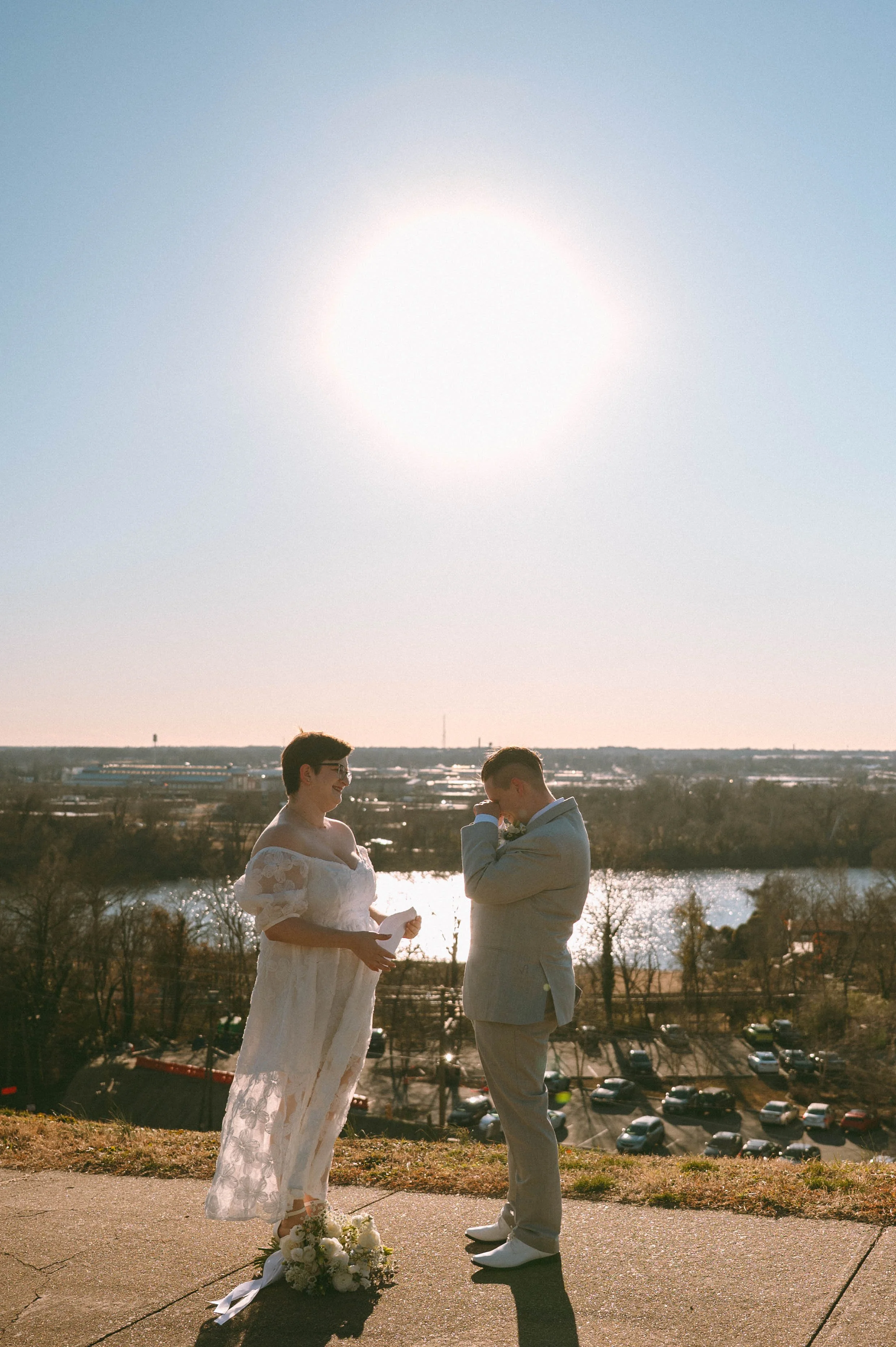 A couple standing outdoors during a sunny day, exchanging vows or rings, with the bride wearing a lace wedding dress and holding a bouquet of flowers, while is dressed in a light-colored suit. A bouquet of flowers is on the ground near the bride's feet.