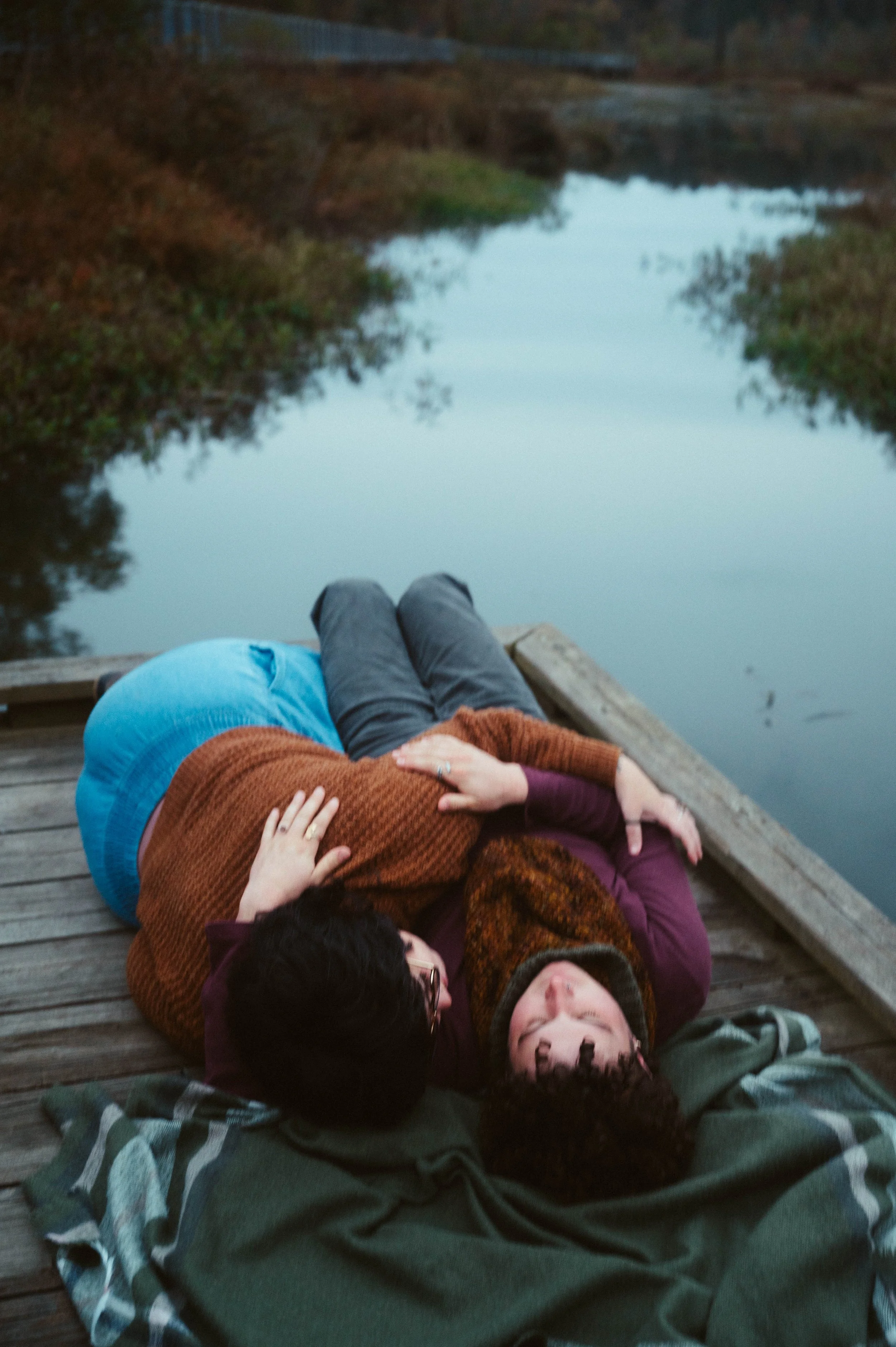 A couple lying on a wooden dock by a calm creek, with their heads together and bodies relaxed, enjoying the outdoor setting at dusk.