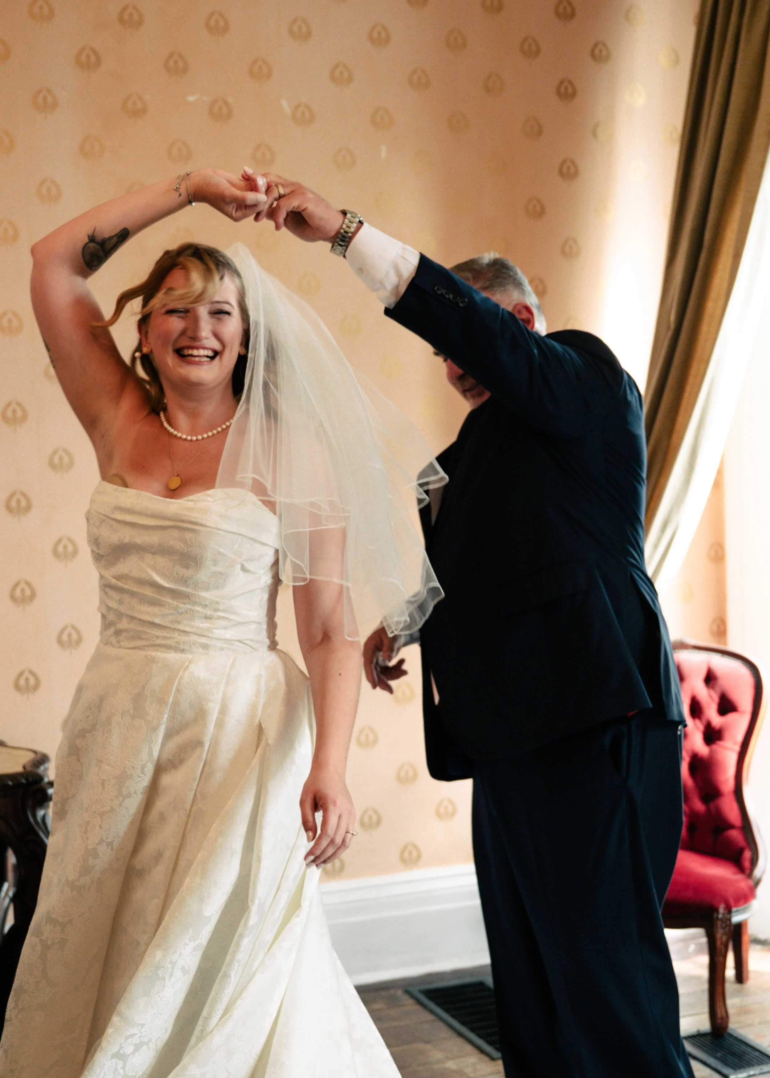 A bride with blonde hair and a big smile, wearing a strapless white wedding dress and a pearl necklace, is spinning with a man, possibly her father, who is wearing a dark suit. They are dancing together at a wedding reception in a room with beige wal