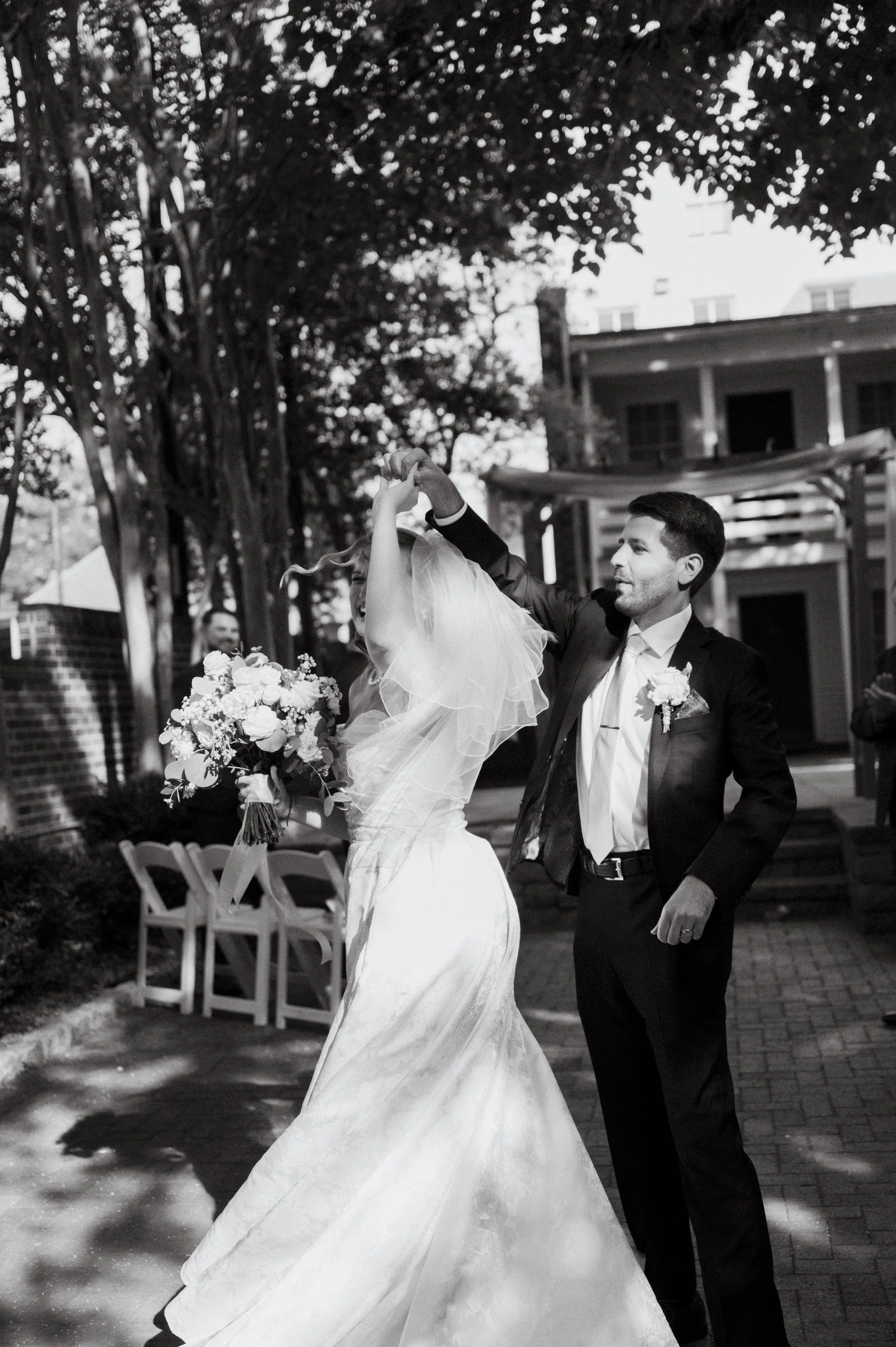 A black and white photo of a newlywed couple dancing outdoors. The man is in a suit and the woman is in a wedding gown holding a bouquet, with trees and a building in the background.