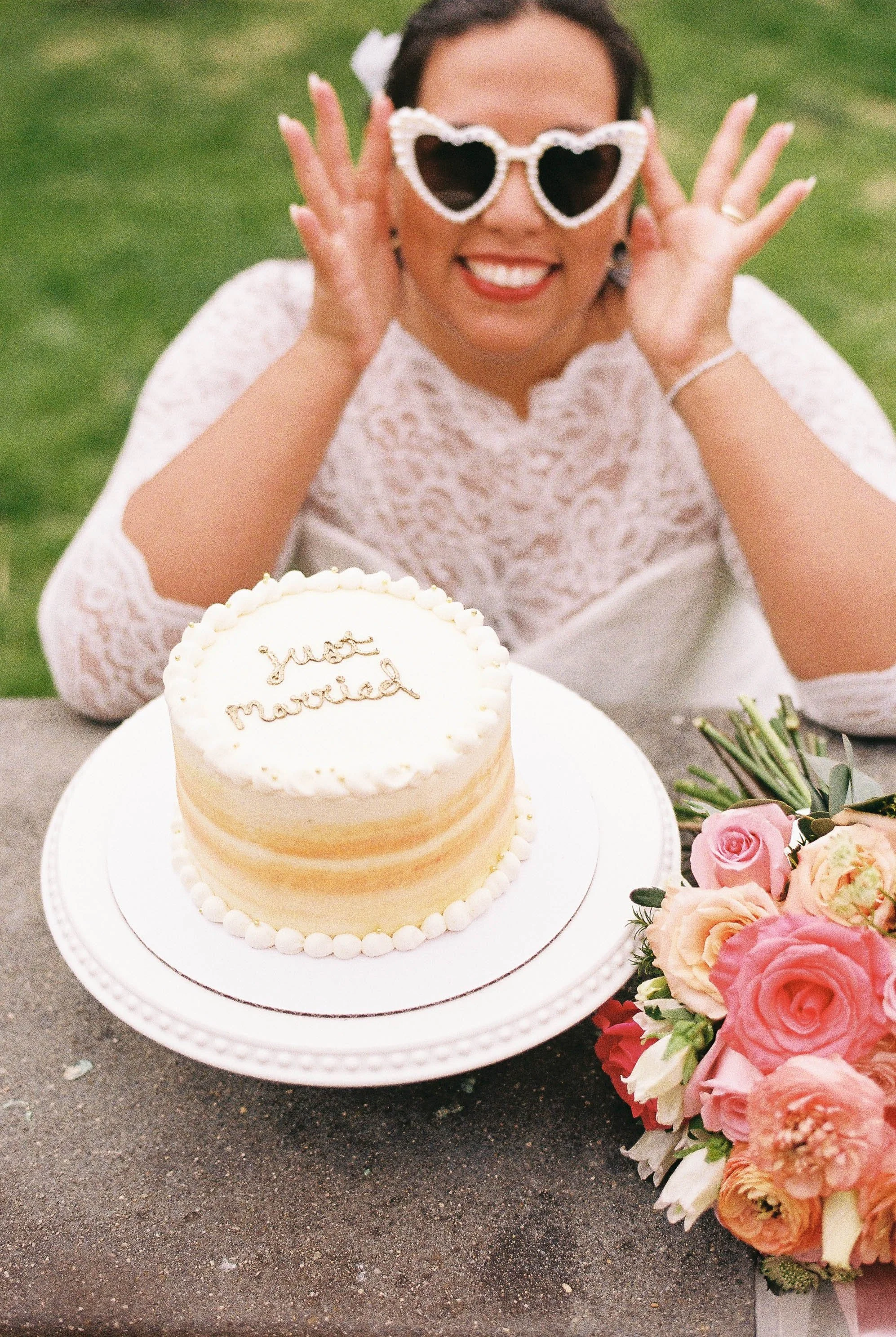 A woman in a white lace dress wearing heart-shaped sunglasses smiling, with a wedding cake that has a message 'just married' and a bouquet of pink and peach roses on a table outdoors.