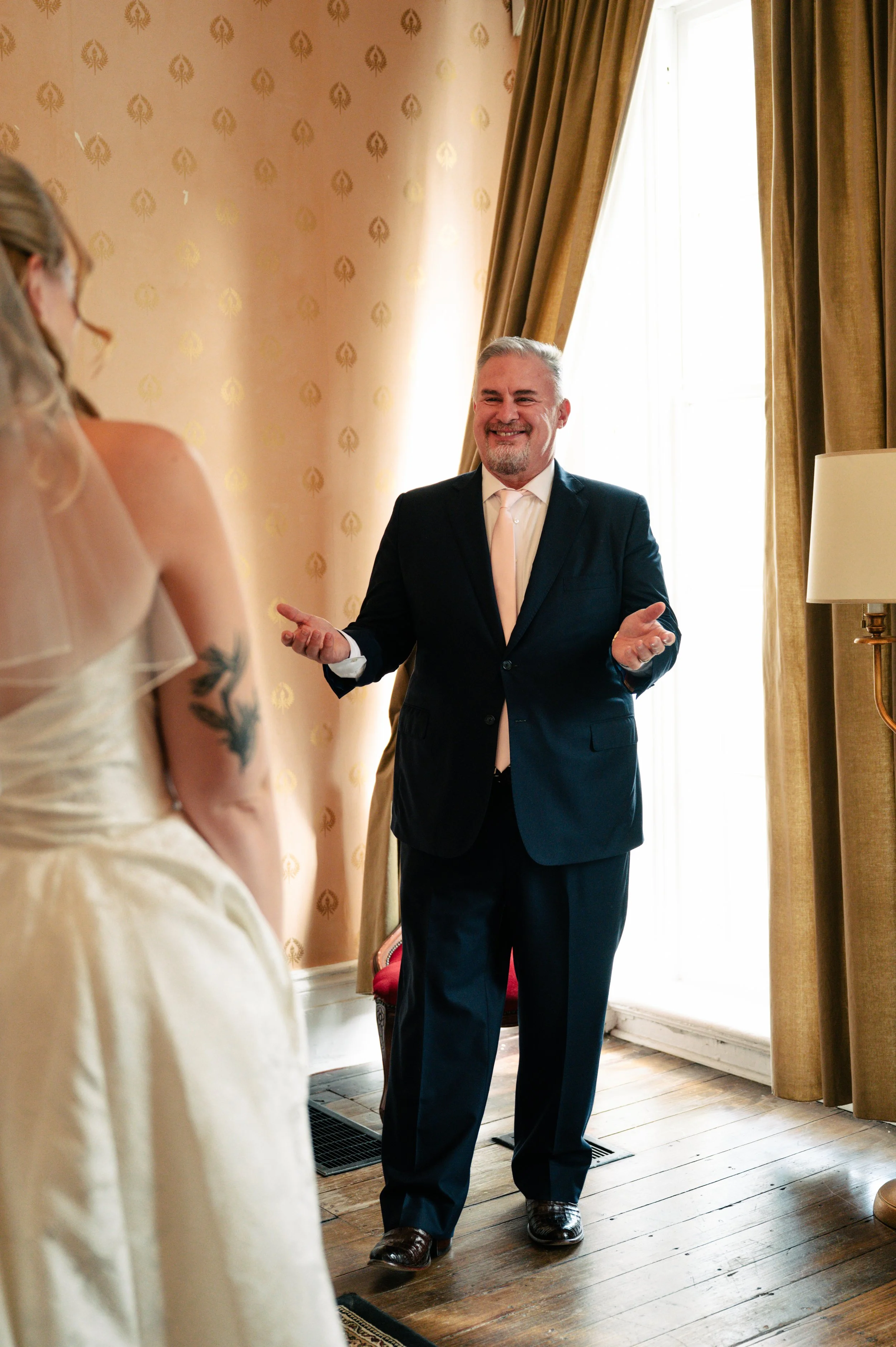 A man in a navy suit, white shirt, and light tie smiling with open hands, standing inside a warmly lit room with large window and curtains, during a special occasion.