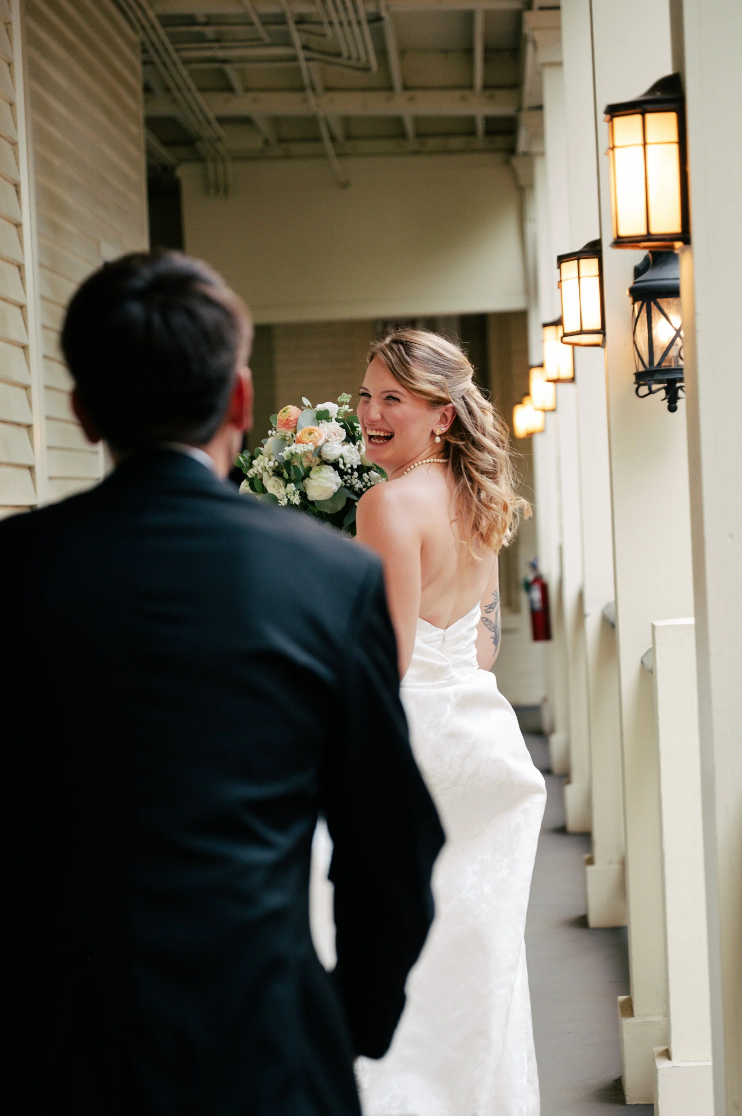 A bride with blonde hair smiling and holding a bouquet of flowers, looking over her shoulder at a man in a black suit, in an indoor setting with yellow exterior lamps on the wall.