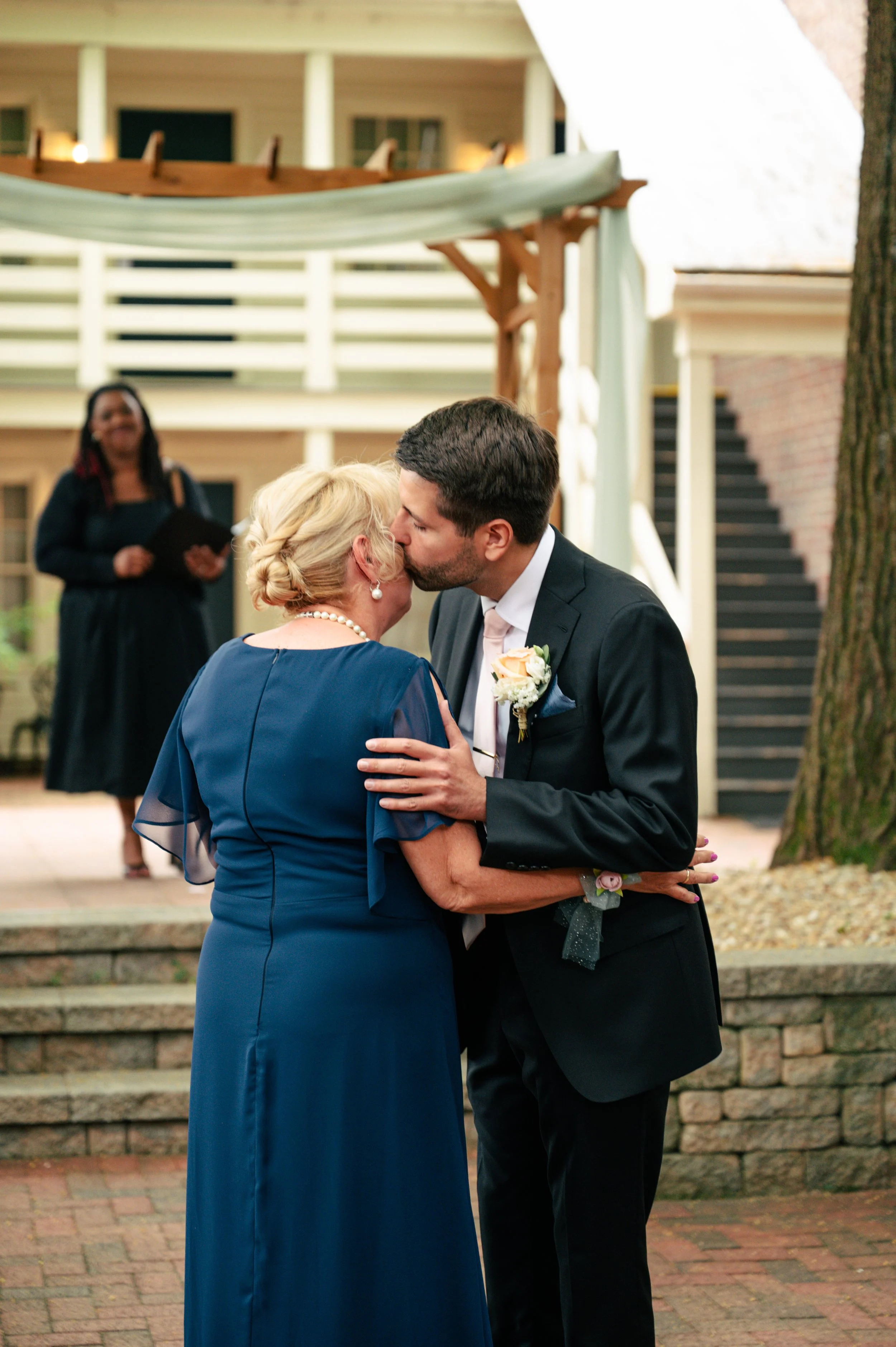A man and woman sharing a kiss during a wedding ceremony outdoors, with a woman in the background holding a book and smiling, and a building with balconies behind them.