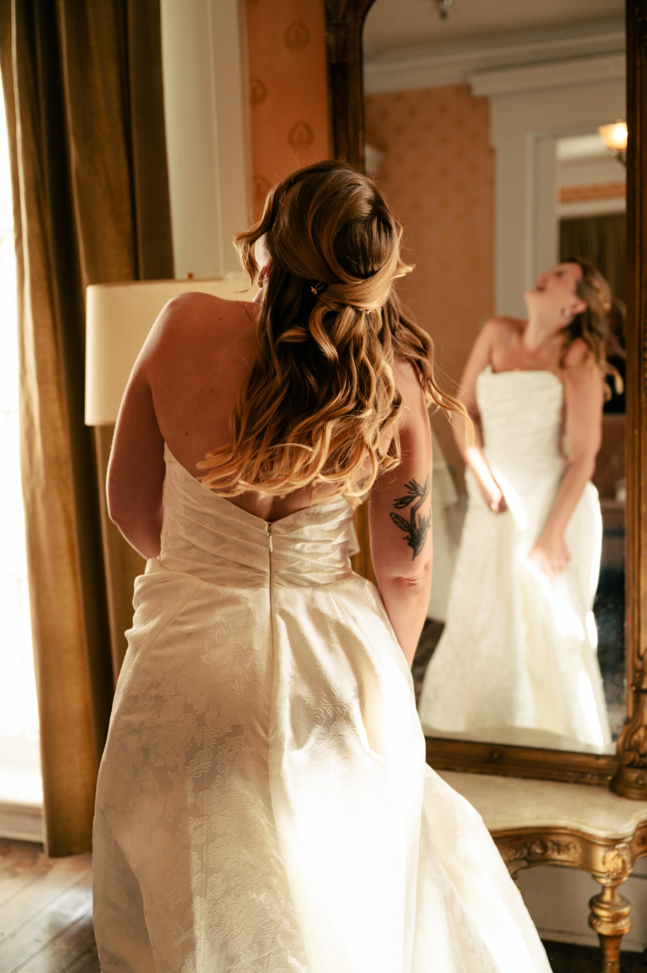 A woman with styled hair and a tattoo on her arm is looking at herself in a large mirror while wearing a strapless white wedding gown with a textured floral pattern.