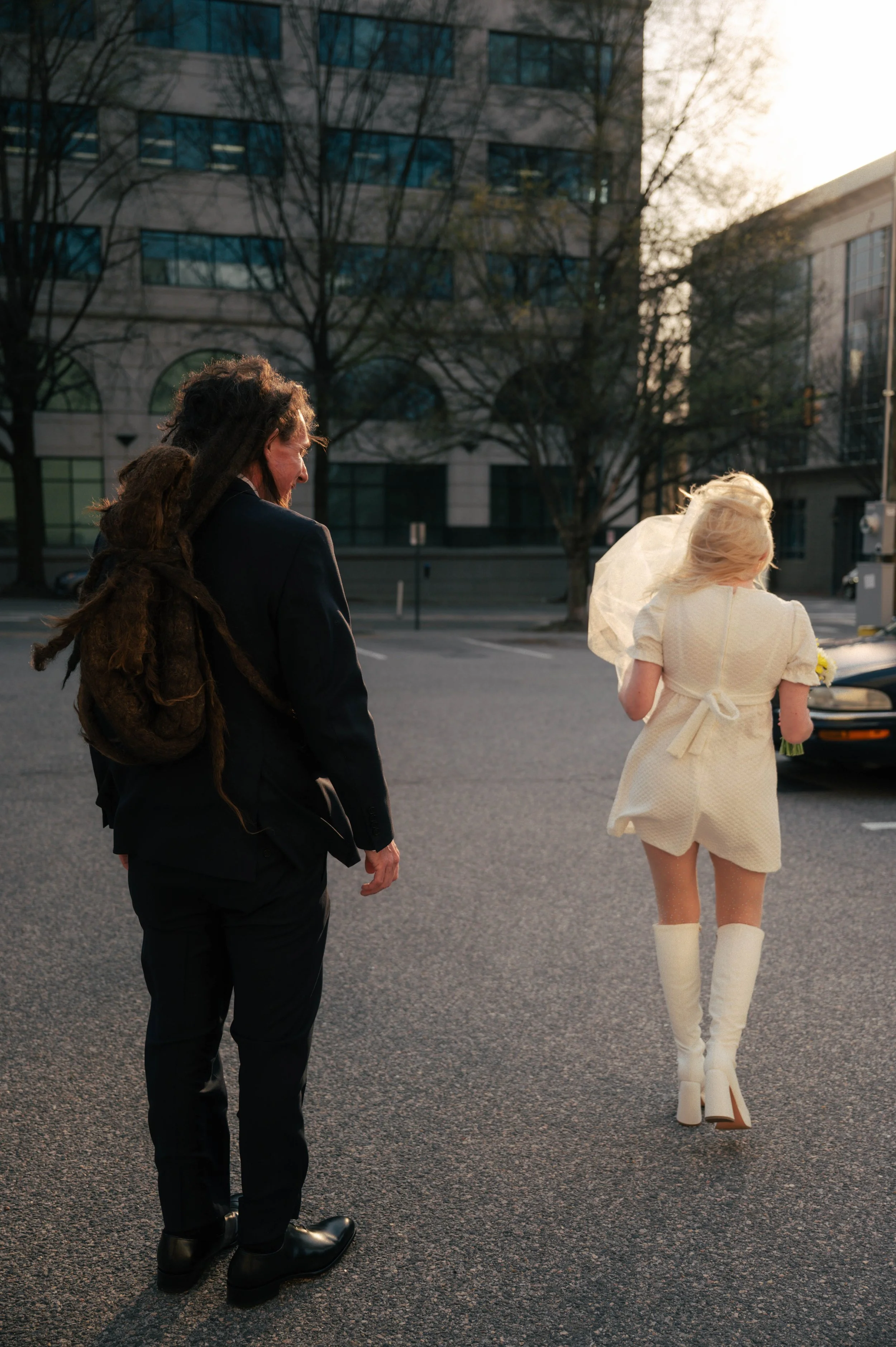 A man with dreadlocks and a woman with long blonde hair, dressed in white, walking on a city street during sunset.