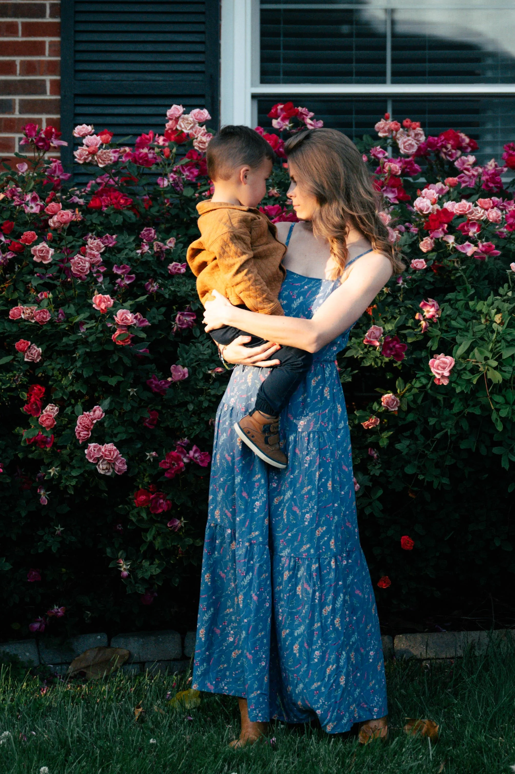 A woman wearing a long blue floral dress holding a young boy in a brown jacket in front of a garden with pink roses. They are standing on grass near a brick house with a window and black shutters.
