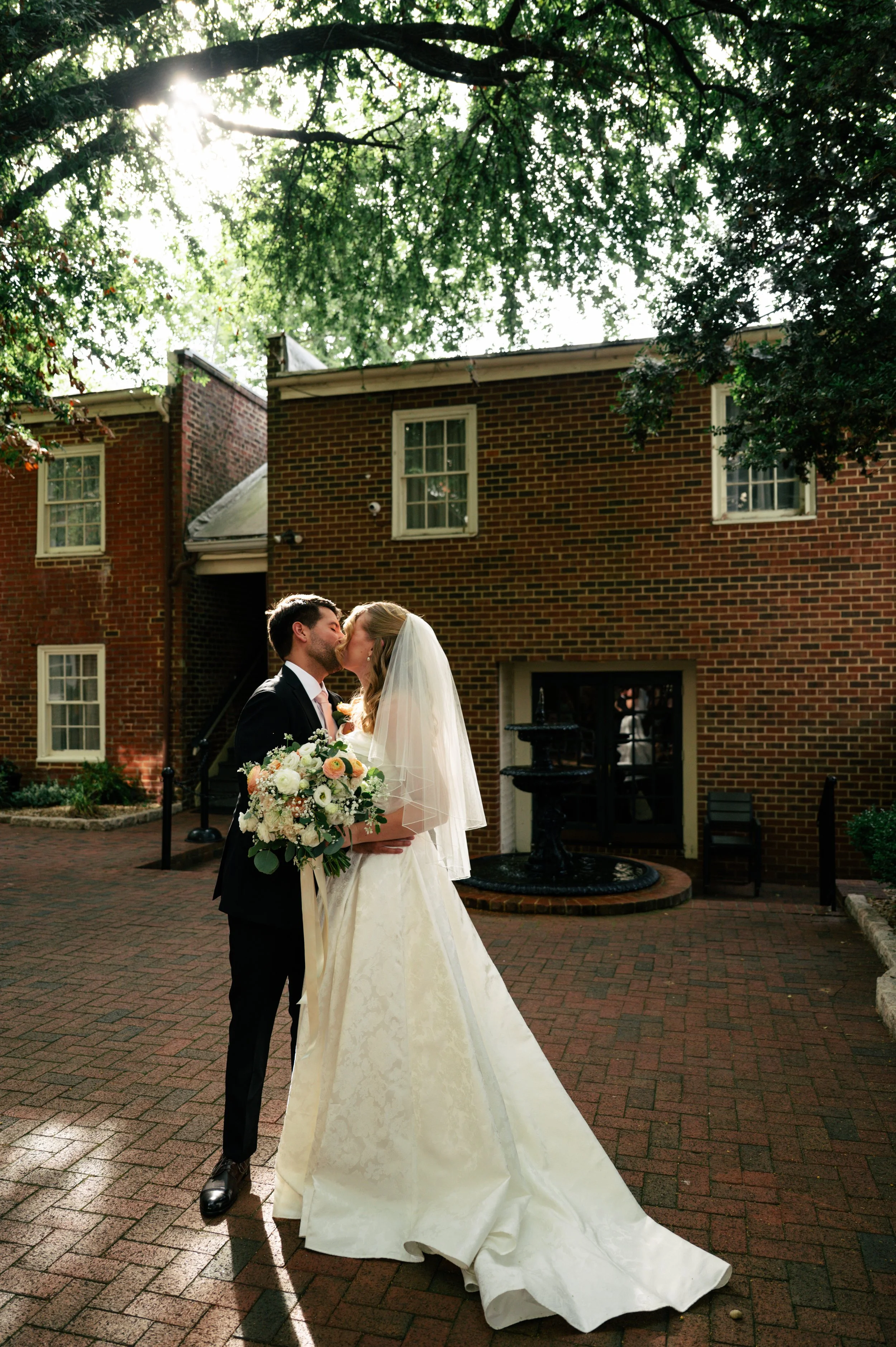 A bride and groom sharing a kiss outdoors in front of a brick building with trees overhead, sunlight filtering through the leaves. The bride is holding a large bouquet, and the groom is dressed in a dark suit. The scene is romantic and serene, typica