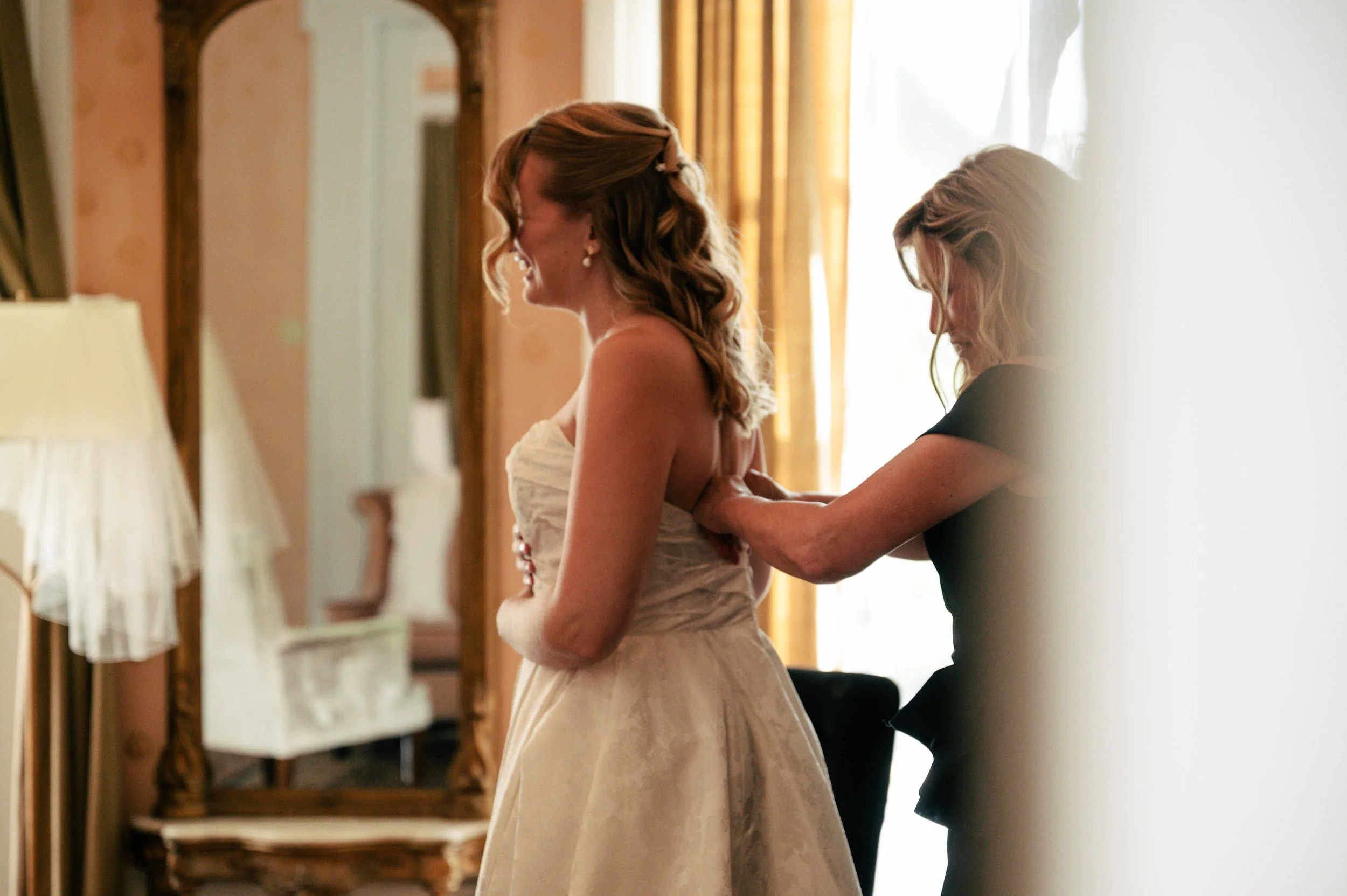 A woman in a wedding dress having her corset tightened from behind by another woman in black. They are in a room with warm lighting and curtains, with a mirror and furniture visible in the background.