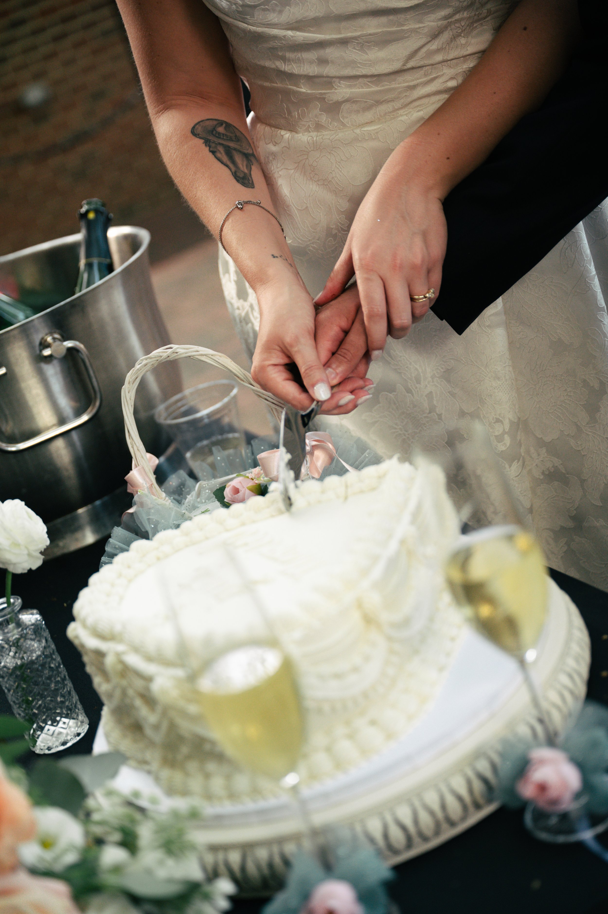 Close-up of a wedding cake with white icing and intricate design, surrounded by flowers and champagne bottles, as a bride and groom cut the cake together.