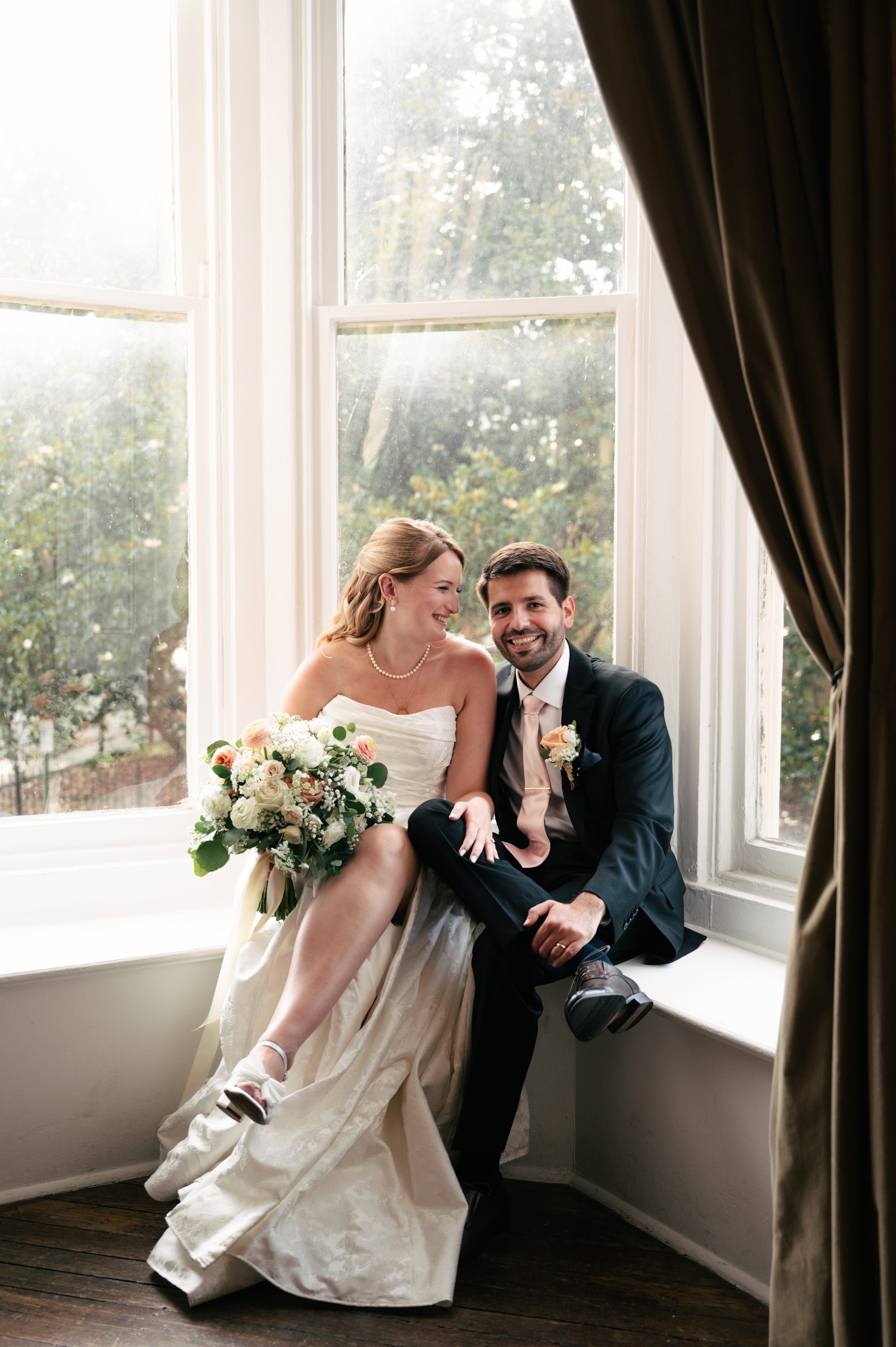 A newlywed couple sitting on a windowsill, smiling, with the bride holding a bouquet and the groom wearing a suit and boutonniere. They are in a room with large windows and dark curtains, with trees outside.
