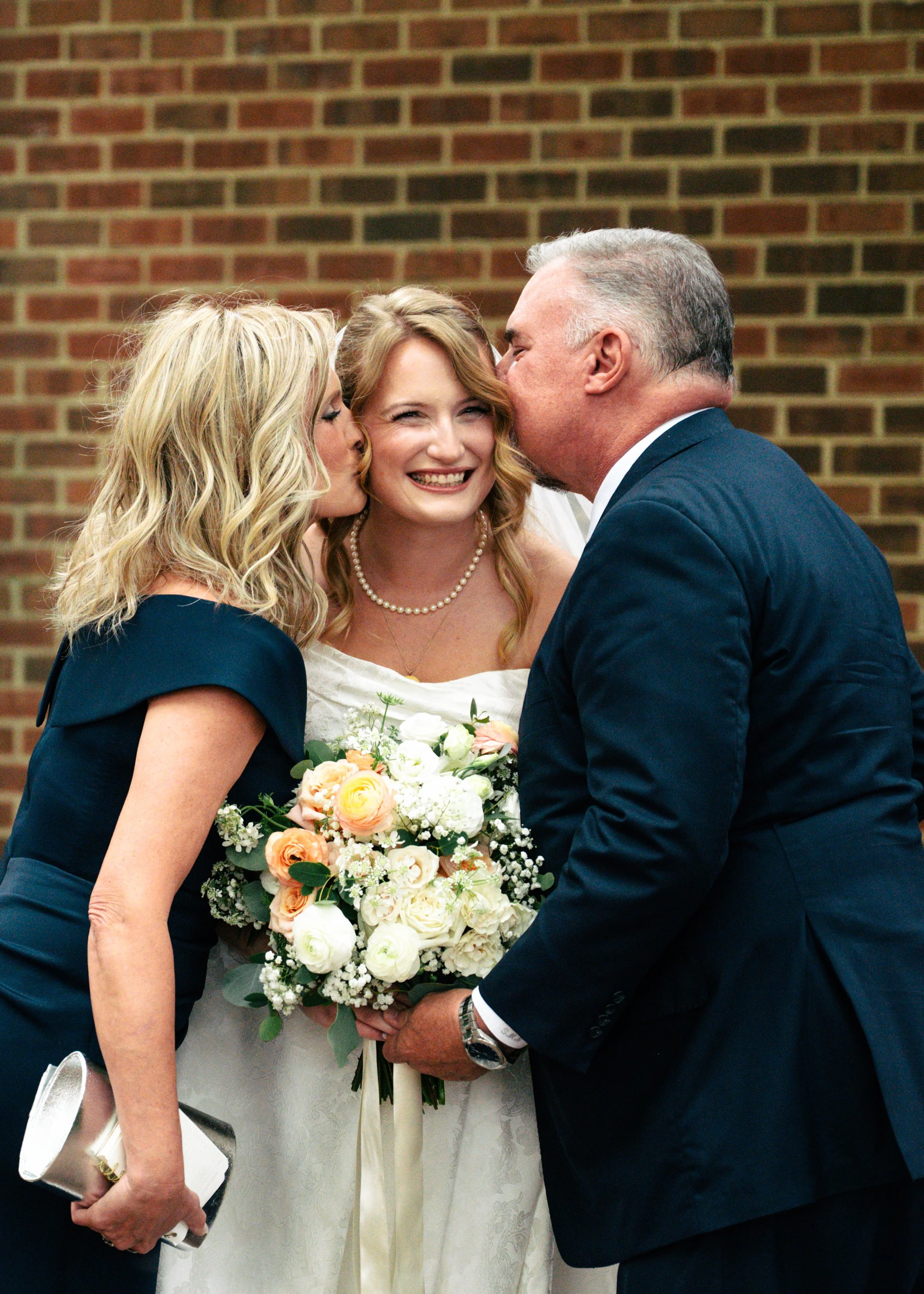 A bride holding a bouquet being kissed on both cheeks by her father and mother during a wedding celebration in front of a brick wall.