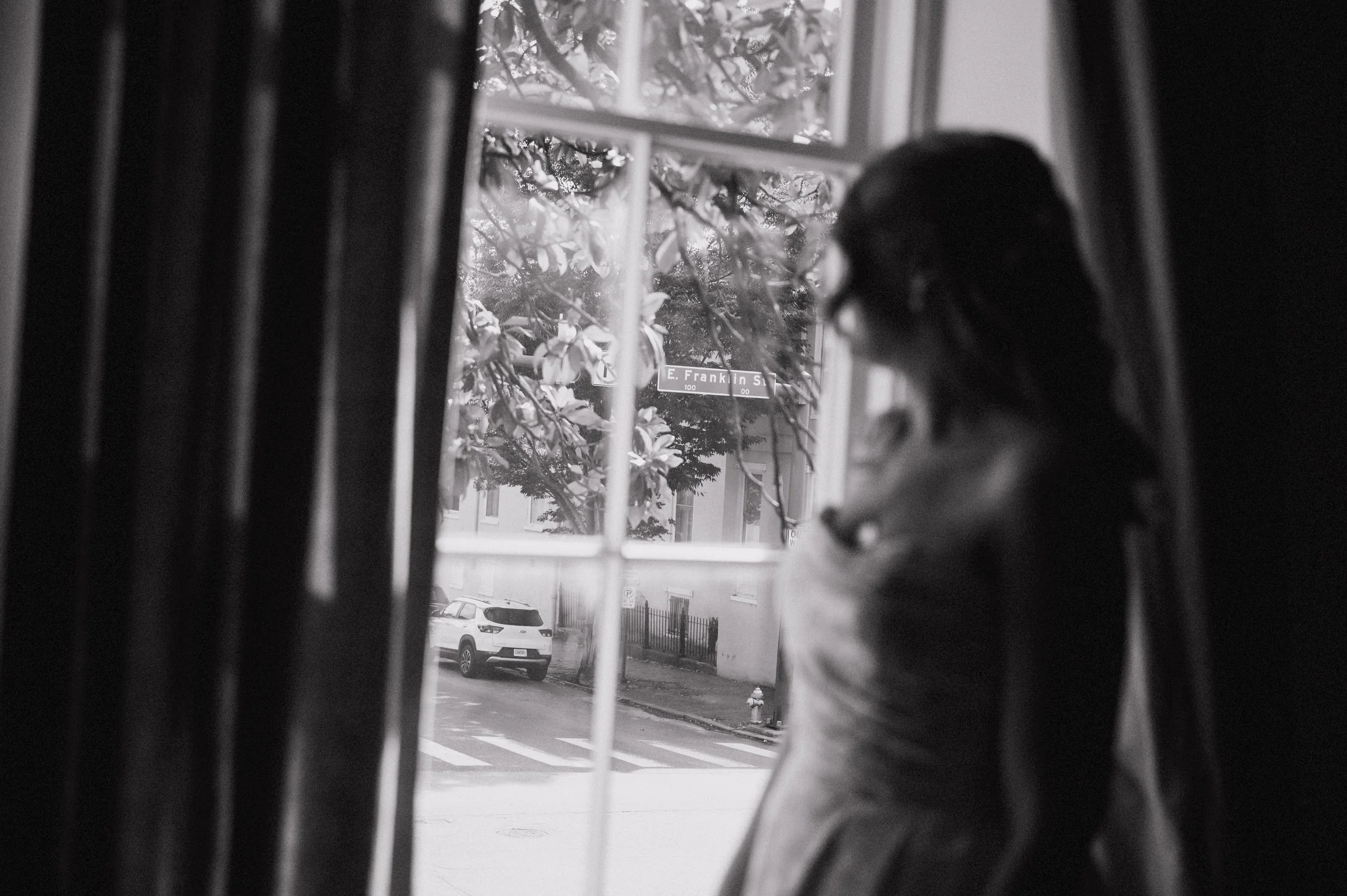 Black and white photograph of a person looking out a window, partially obscured by window bars, with a street scene outside showing parked cars, a tree, and a street sign that reads 'E. Franklin St'.