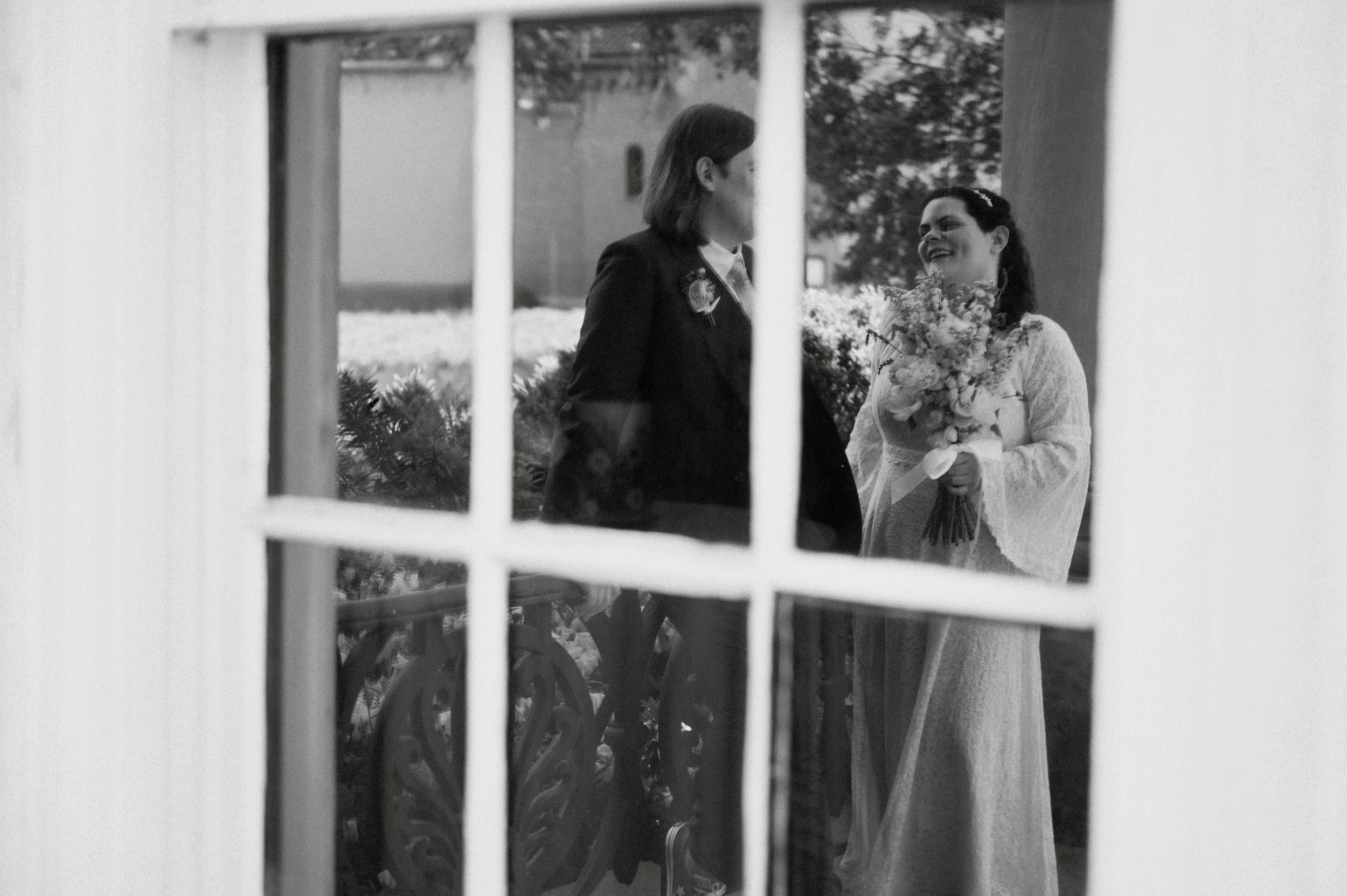 A woman in a white wedding dress holding a bouquet, smiling at a man in a dark suit, seen through a window with multiple panes.