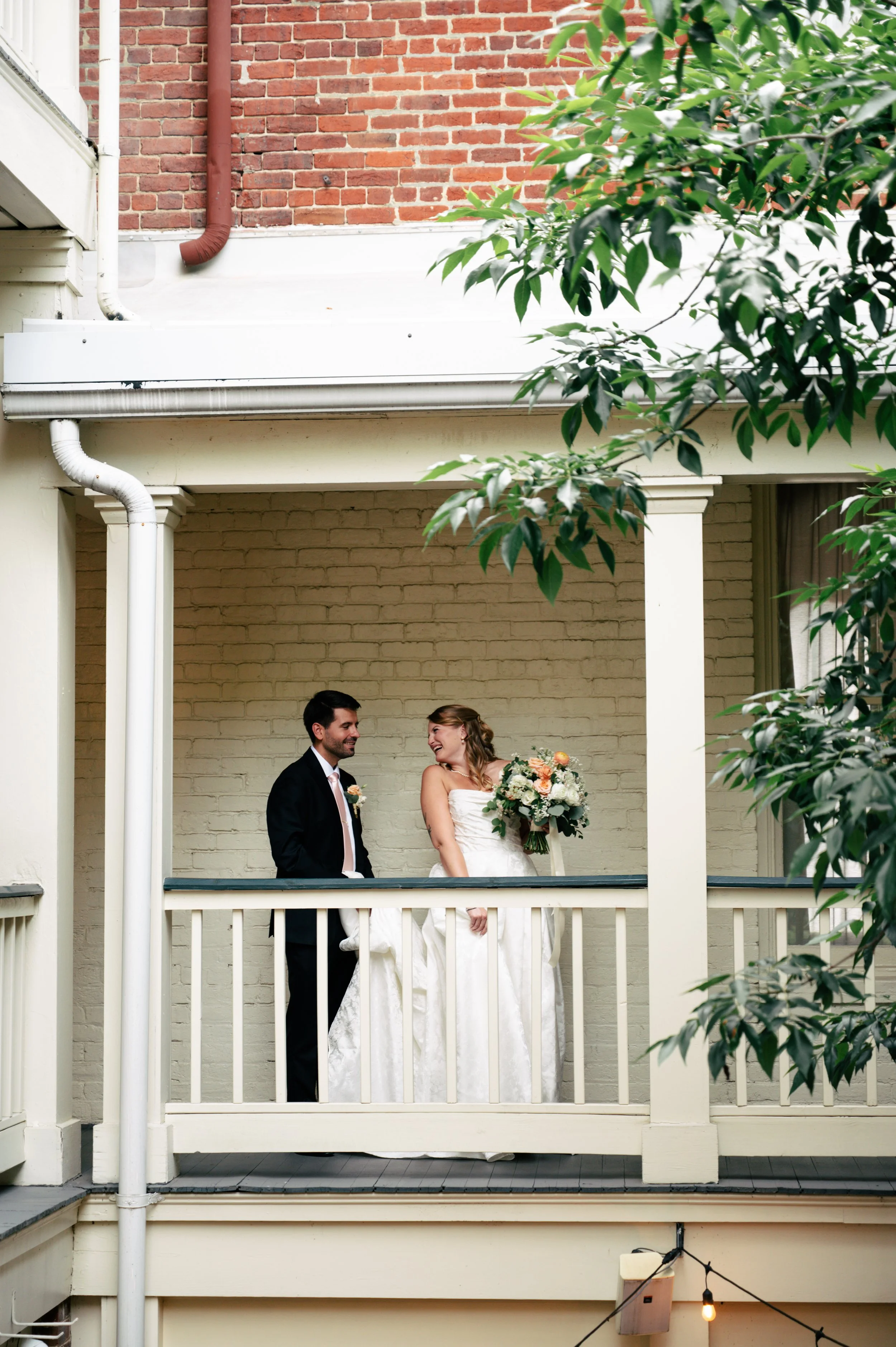 A bride and groom standing on a small balcony, smiling and looking at each other. The bride holds a bouquet and is wearing a white wedding dress. The groom is dressed in a black suit with a white shirt and peach-colored tie. The background shows a br