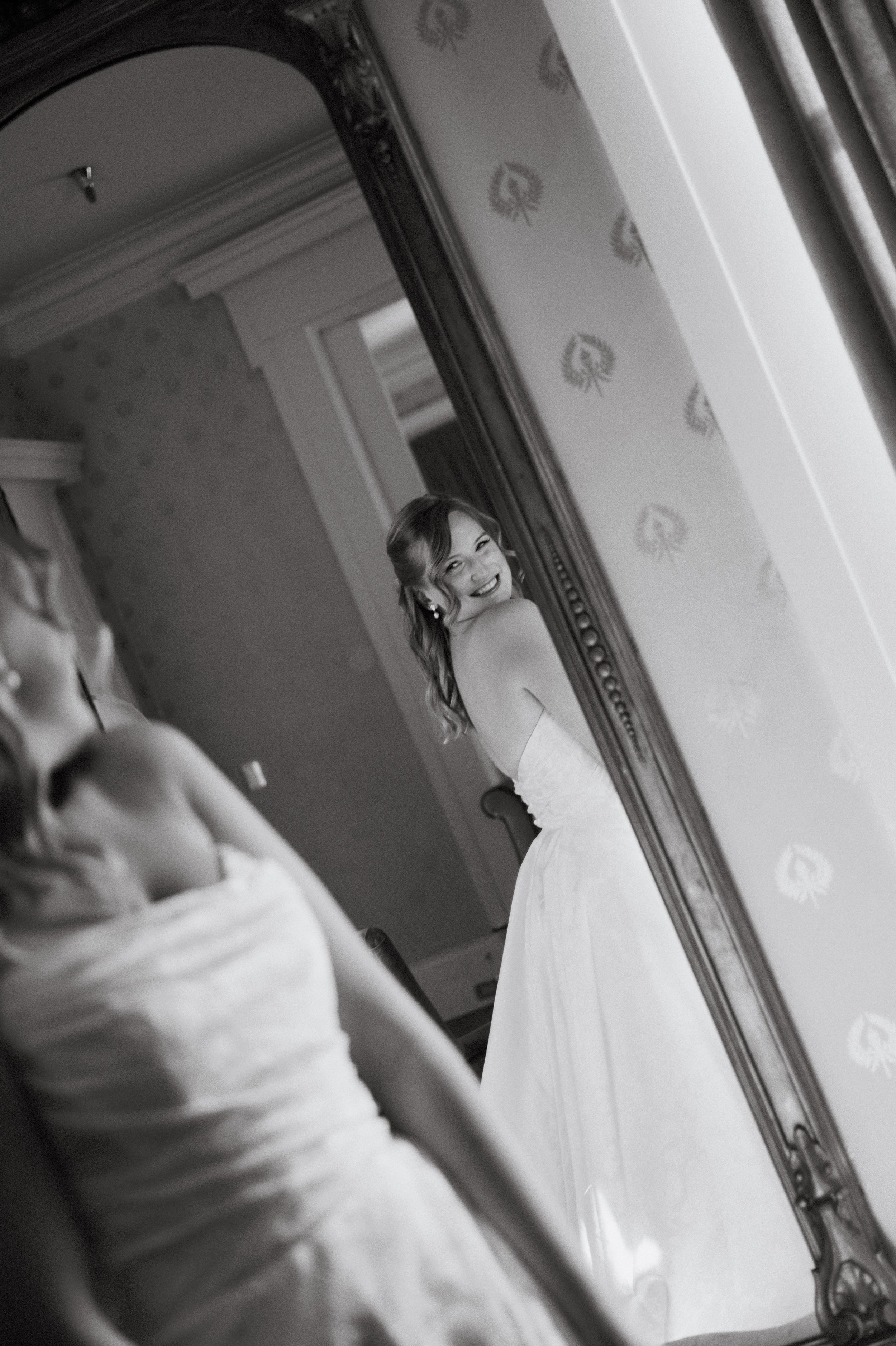 A woman in a wedding dress smiling while looking into a mirror in a room with patterned wallpaper.
